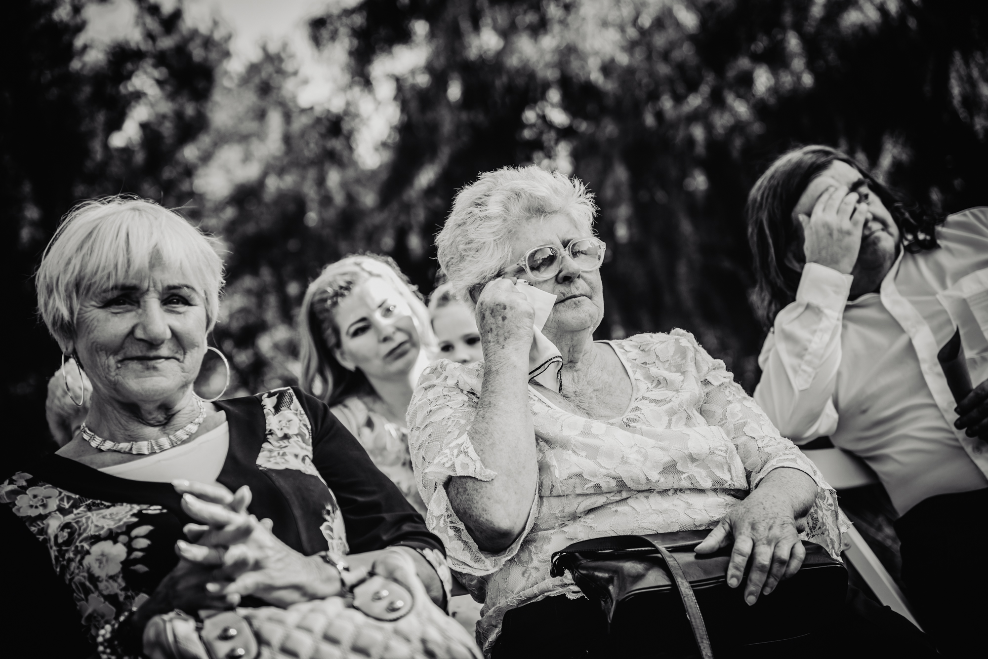 Elderly wedding guest wipes away tears during an emotional outdoor ceremony.