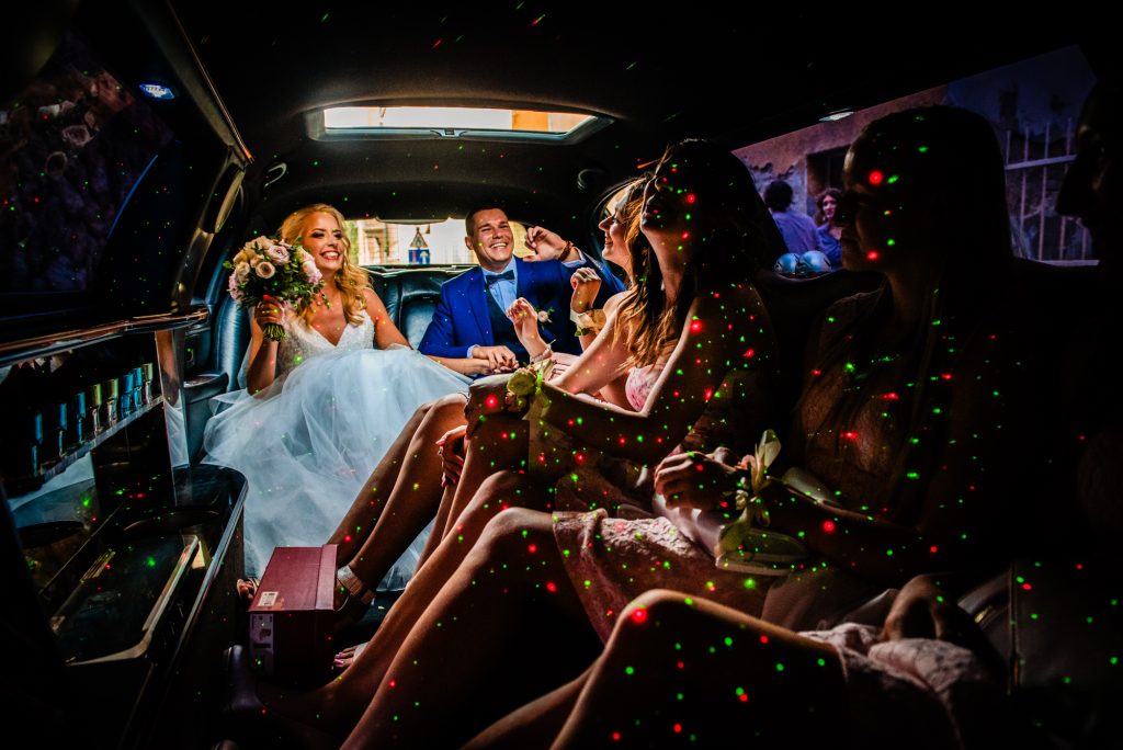 Bride and wedding party laughing inside a limousine lit with colourful party lights in Budapest.