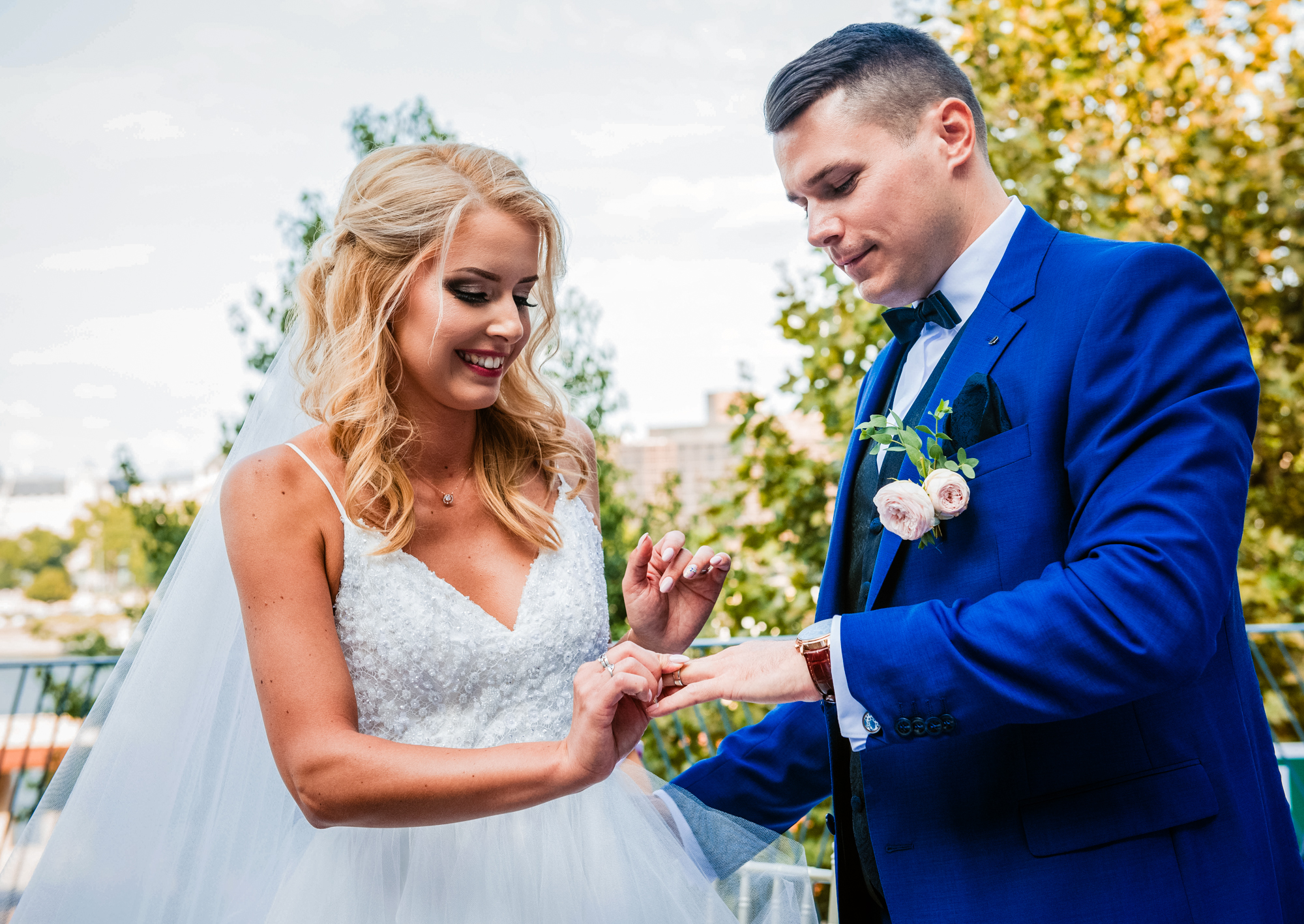 Groom placing wedding ring on bride’s finger during outdoor Budapest wedding ceremony with blue suit and veil.