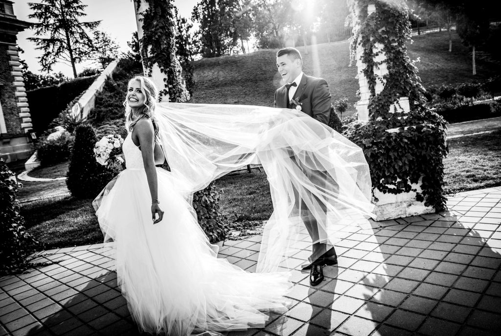 Groom holding bride’s veil while laughing during playful black and white wedding portrait outdoors.