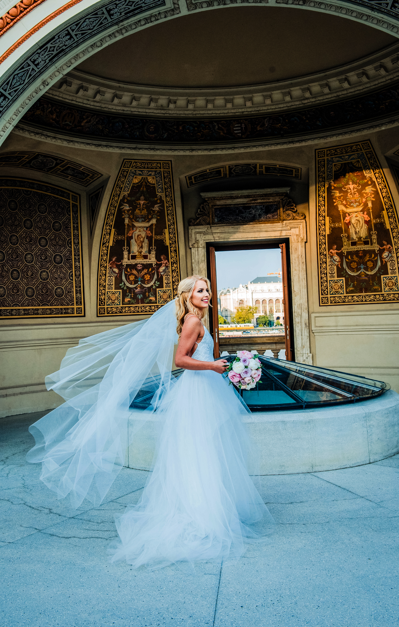 Bride posing with flowing veil beneath ornate historic pavilion during Budapest wedding portrait session.