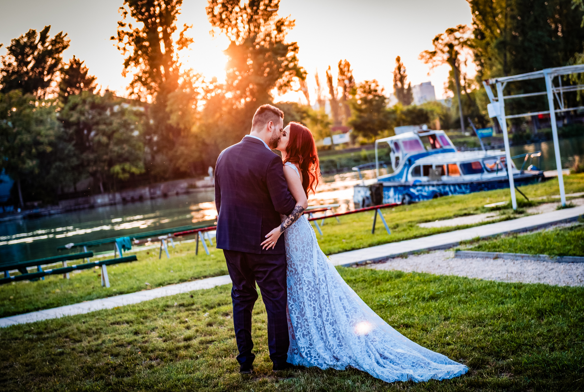 Newlyweds kiss beside the Danube during golden hour near moored boats and riverside greenery.