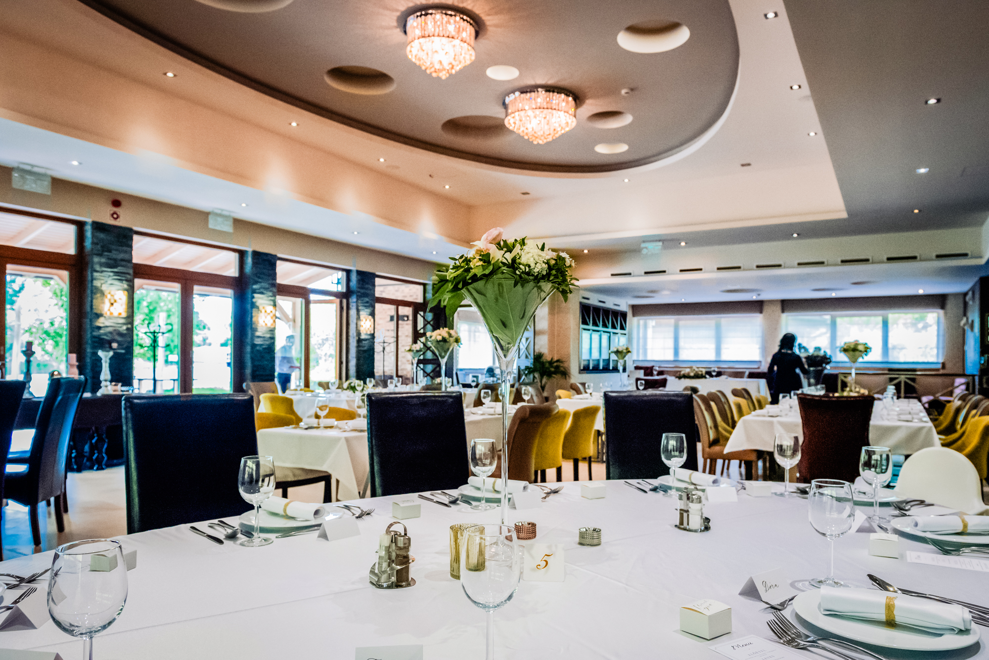 Wide view of wedding reception hall with floral centrepieces and modern chandelier lighting in Budapest.