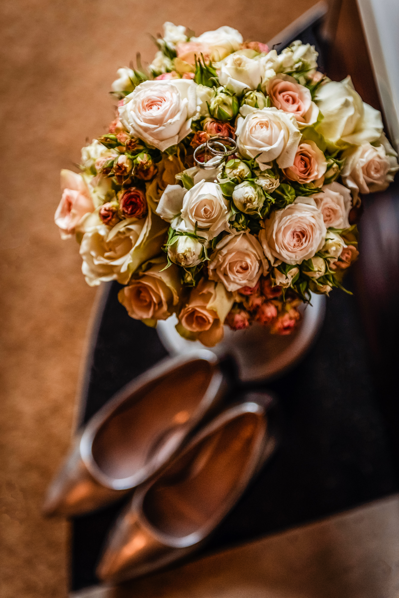 Bridal bouquet with pale roses and wedding rings beside silver bridal shoes on hotel table.