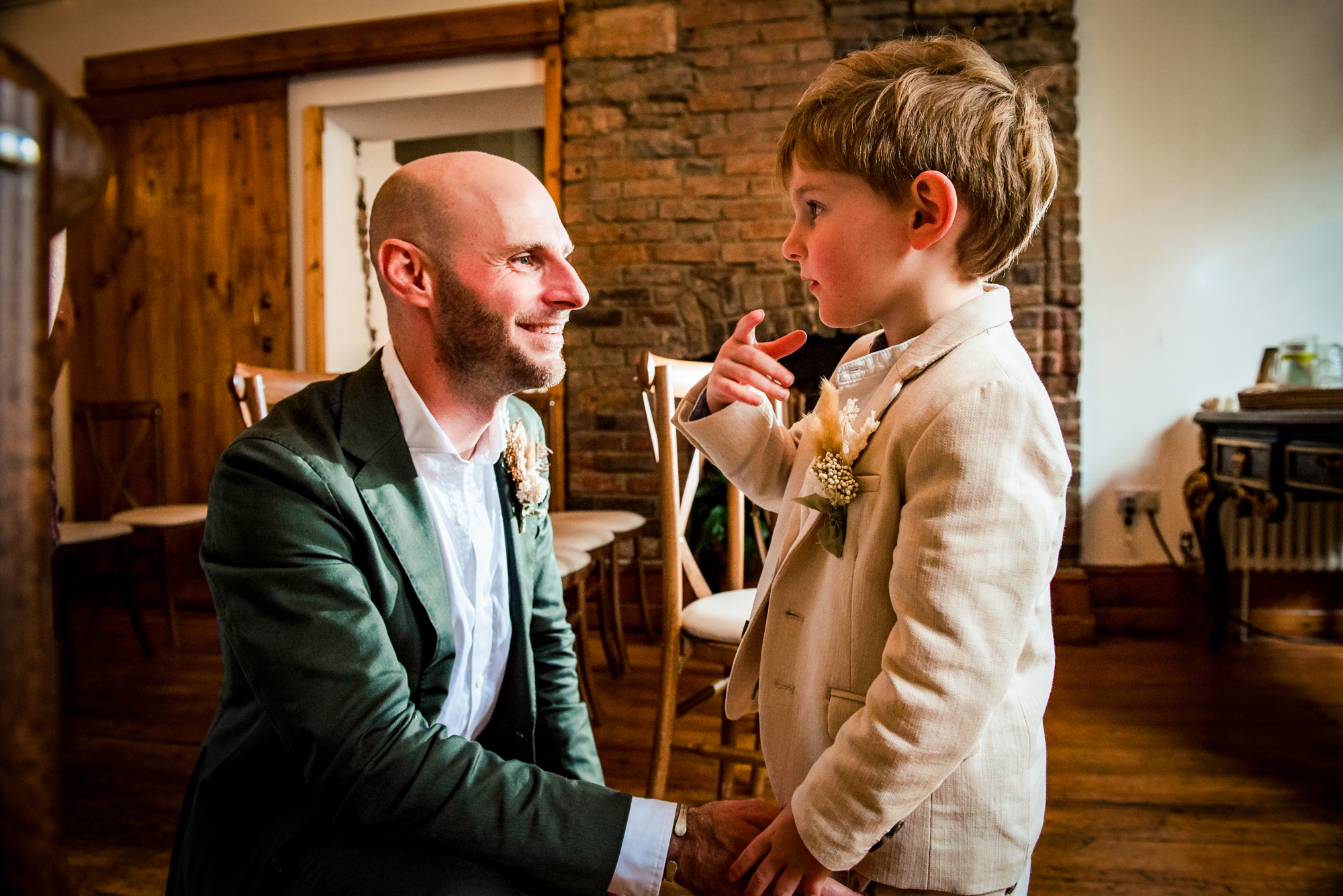 Groom kneeling and smiling at young boy during relaxed indoor wedding ceremony.