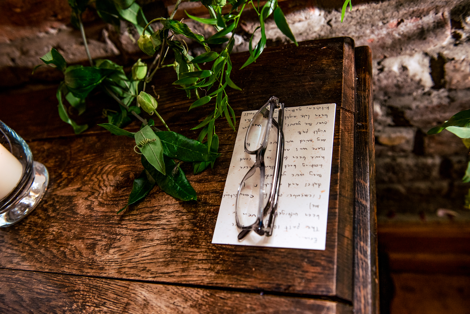 Handwritten vows card and glasses resting on wooden table with greenery details.