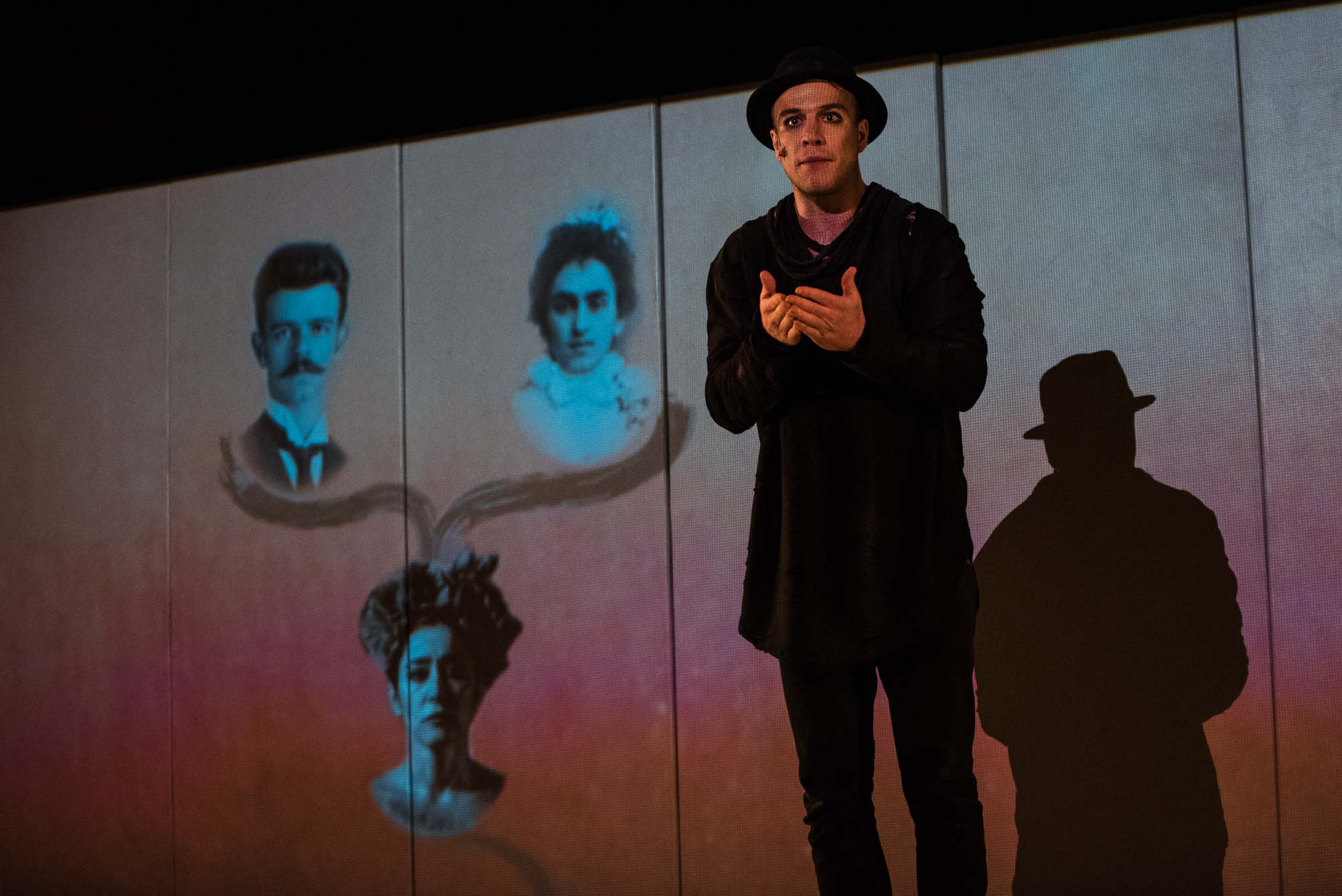 Performer in black costume stands before projected family portraits during Frida Kahlo theatre performance.