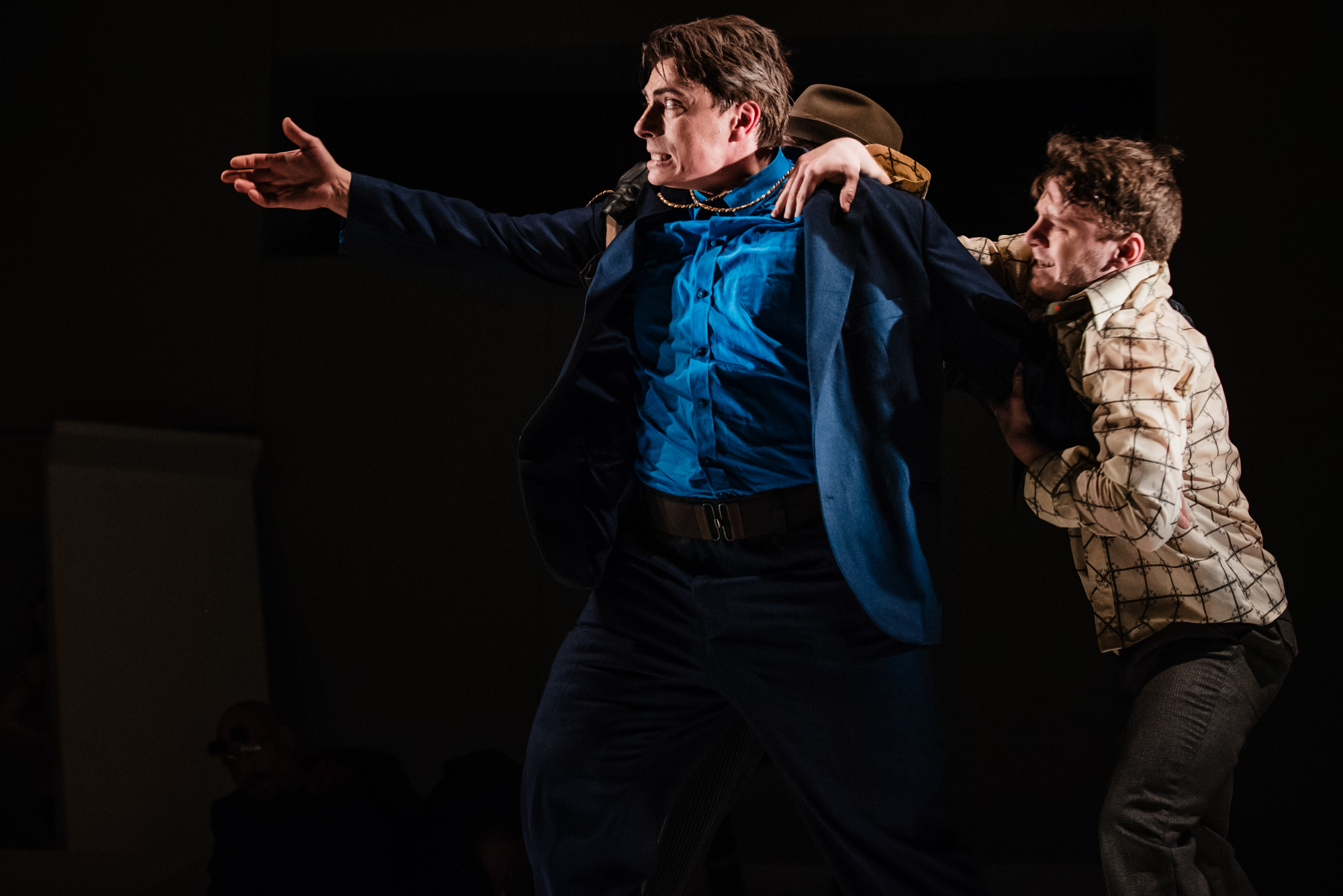 Actor in blue suit gestures dramatically while another performer restrains him on darkened stage.