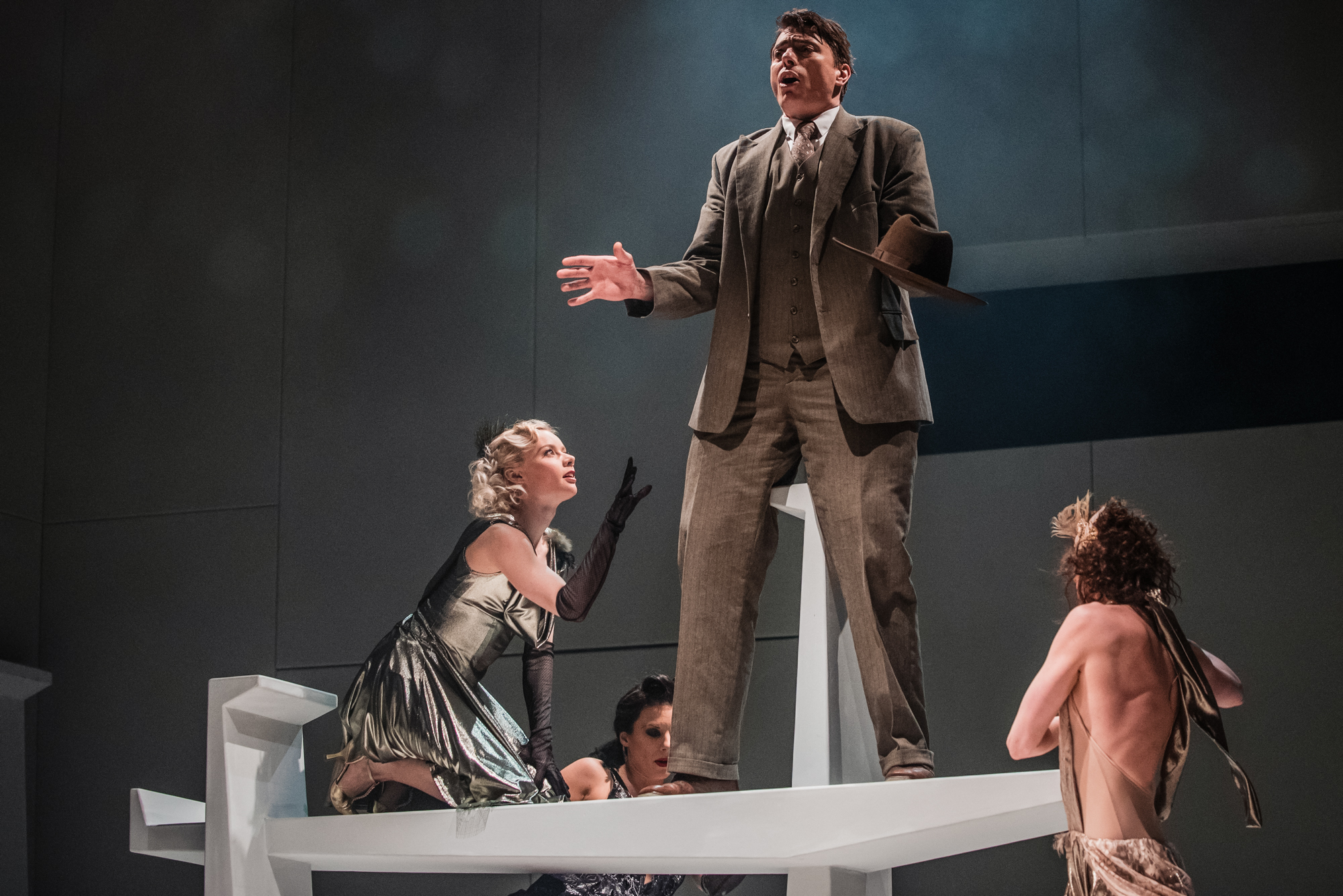 Actor in grey suit stands on elevated stage platform surrounded by dancers during theatre performance.