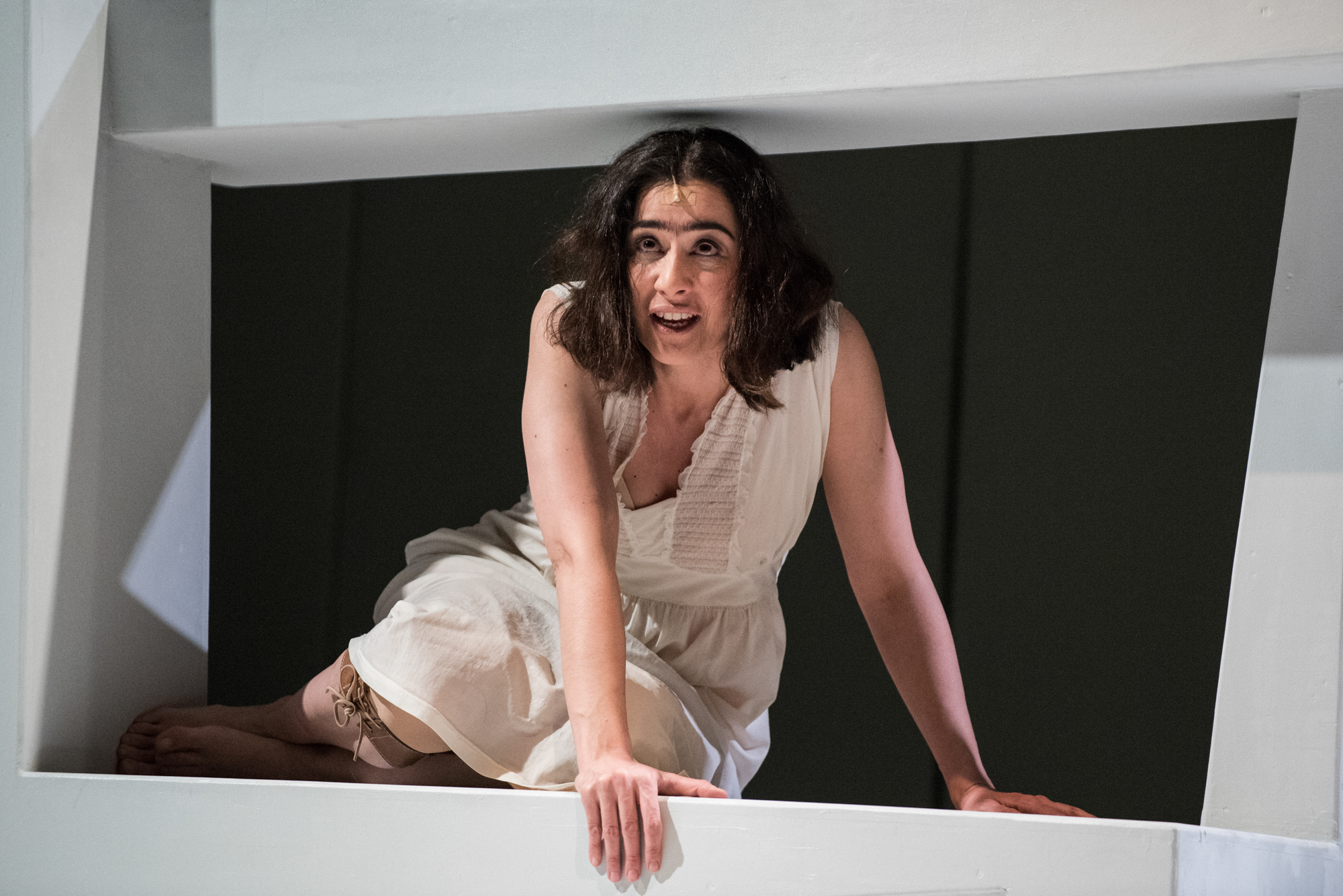 Actress in white dress crawls through geometric stage structure during dramatic theatre performance.