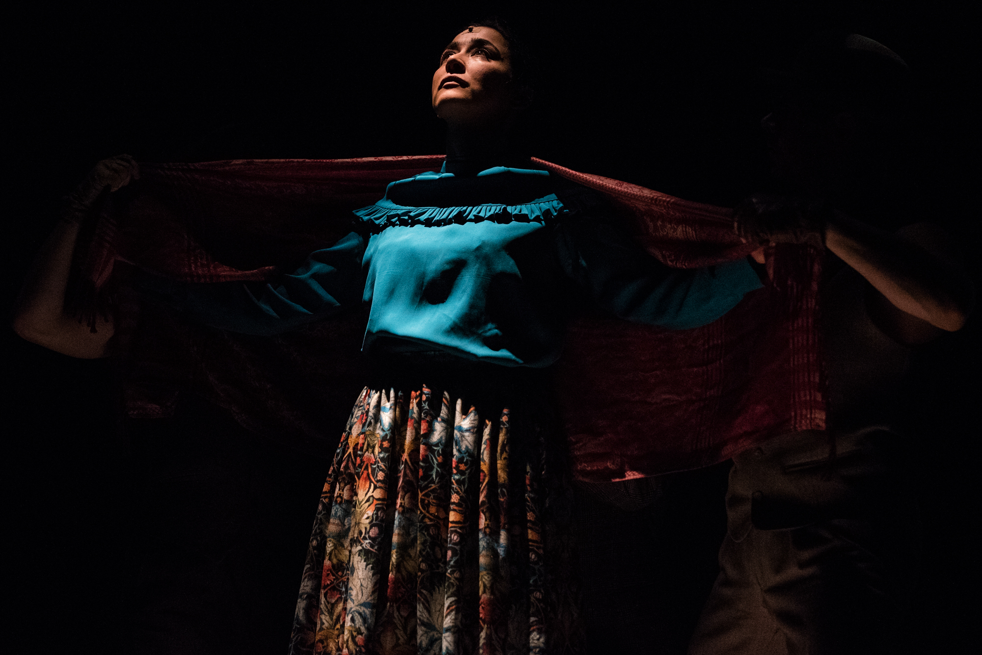 Actress in turquoise blouse stands beneath red shawl in low-lit Frida Kahlo inspired stage scene.