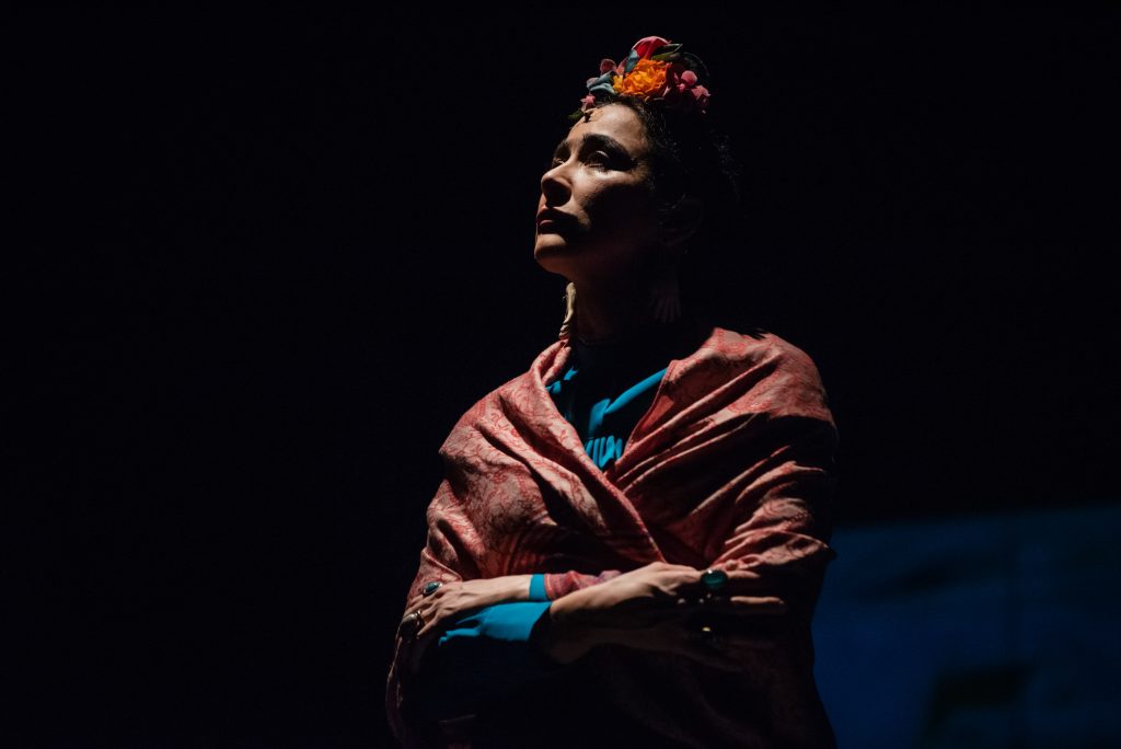 Performer wearing floral crown and pink shawl stands in spotlight during Frida Kahlo theatre production.