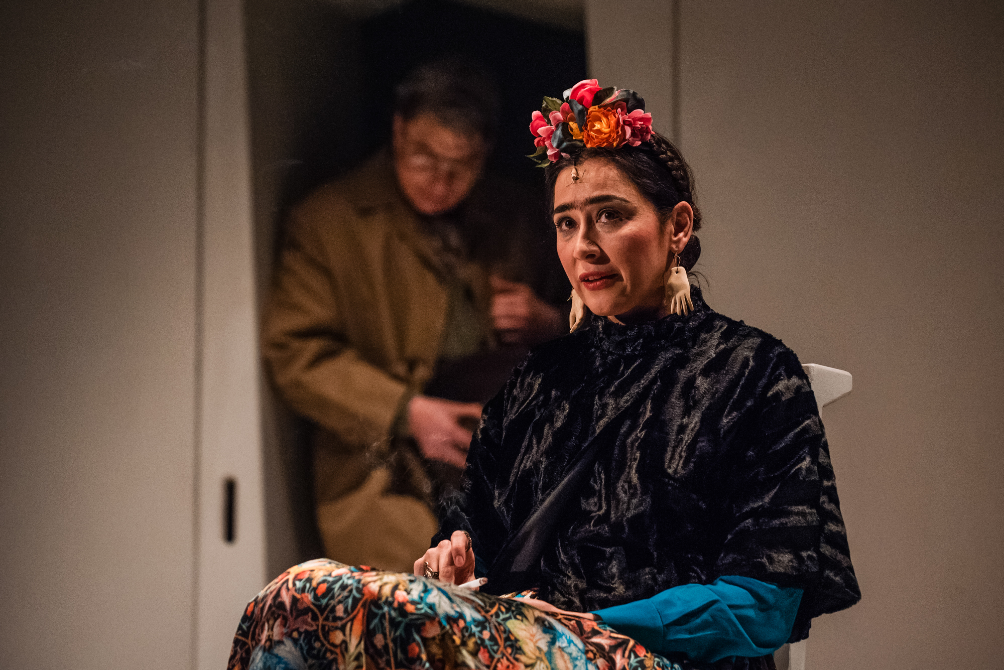Actress portraying Frida Kahlo sits in patterned costume during intimate theatre scene on stage.
