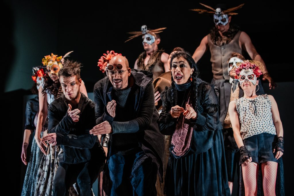 Theatre cast in skull masks and dark costumes perform ensemble scene under stage lighting.