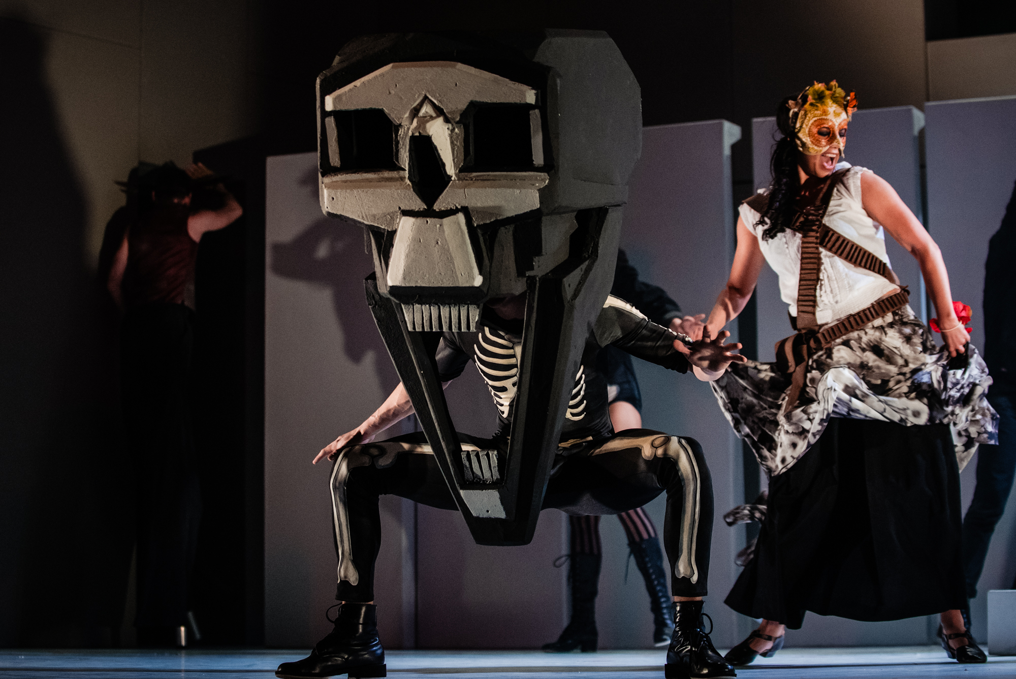 Performer wearing oversized skull head crouches on stage beside masked dancer in floral dress.