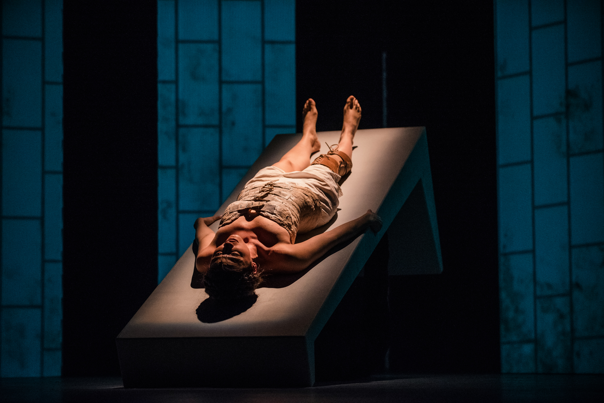 Performer lies on angled stage platform beneath blue-lit backdrop during emotional theatre scene.