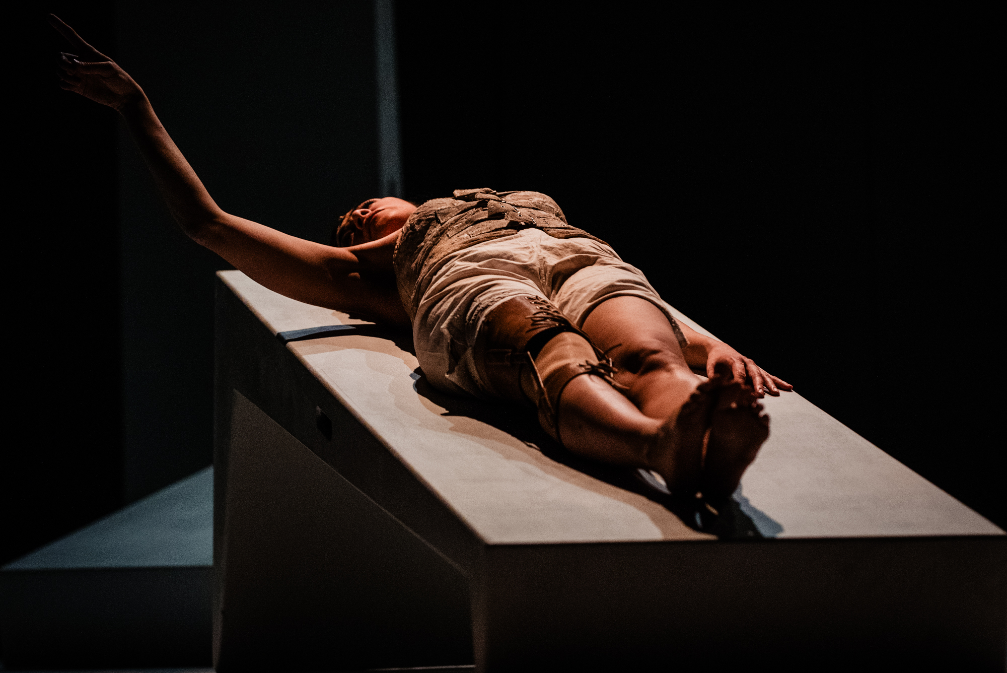 Close-up of performer reaching upward while lying on stage platform in dark theatrical lighting.