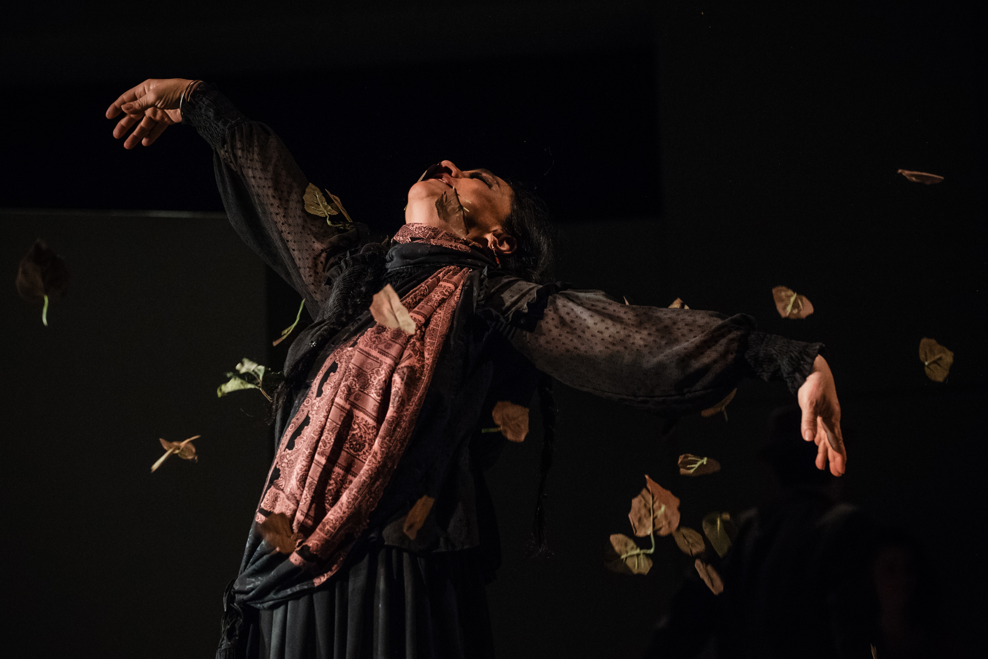 Actress in flowing black costume performs beneath falling leaves during dramatic stage performance.