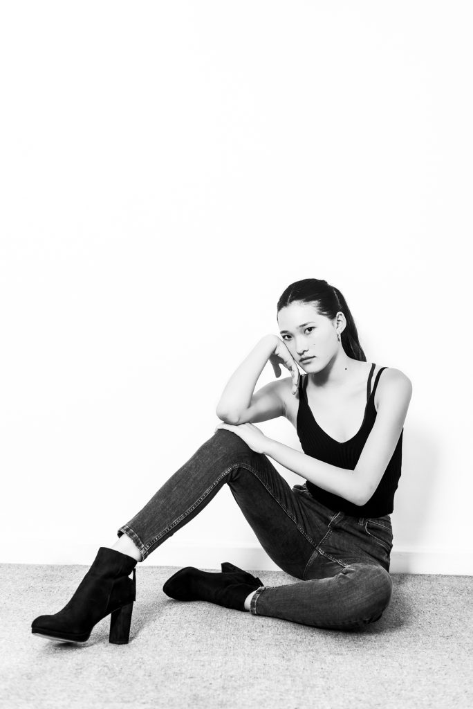Young model seated on carpet floor in monochrome portrait with relaxed editorial pose.