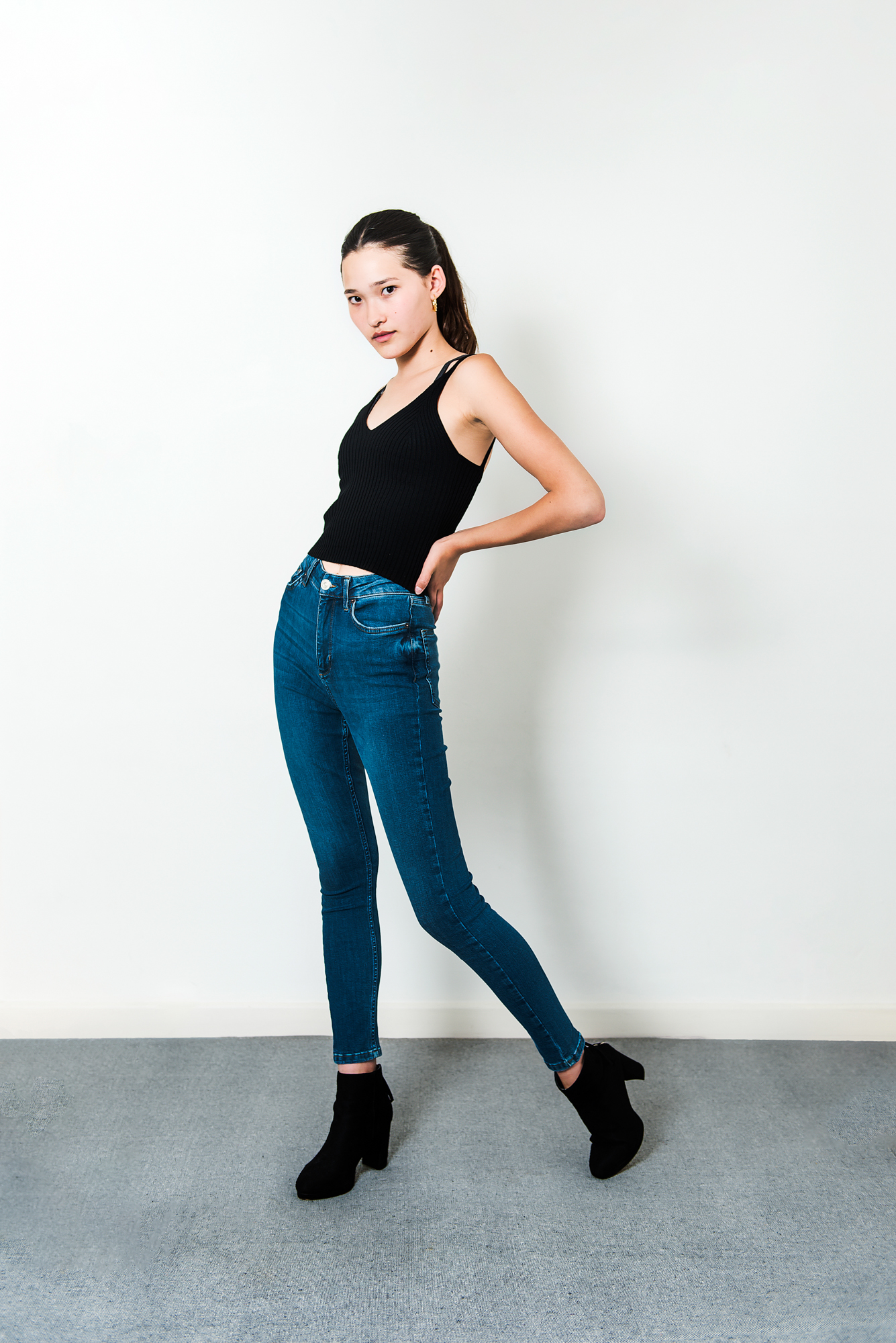 Young model posing sideways in blue jeans and black heels during indoor portrait photography session.