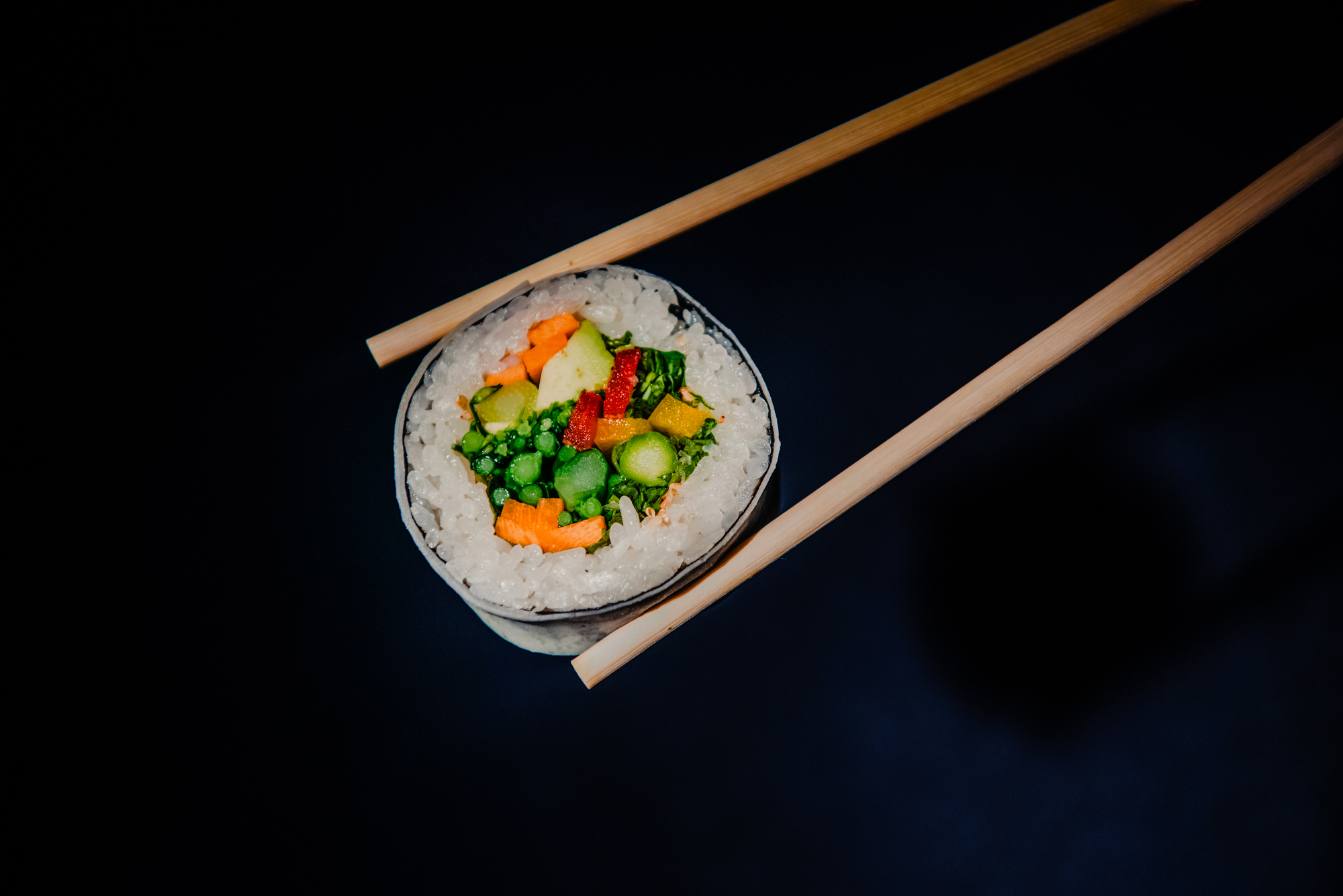 Vegetable sushi roll balanced between chopsticks against a dark studio backdrop.