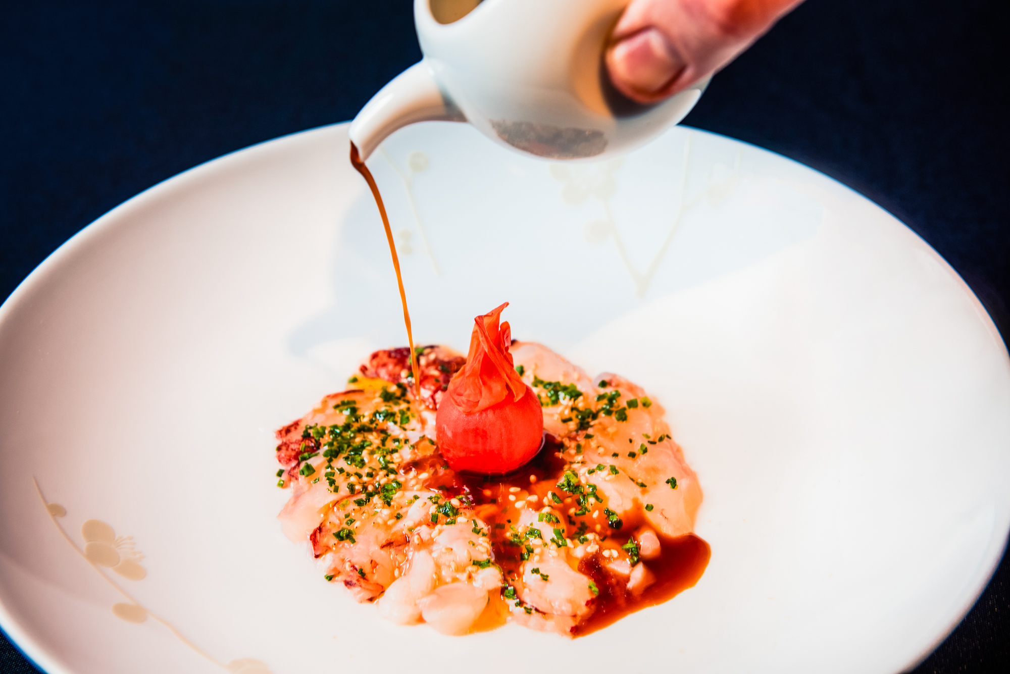 Soy-based sauce being poured over octopus carpaccio with herbs and sesame seeds.