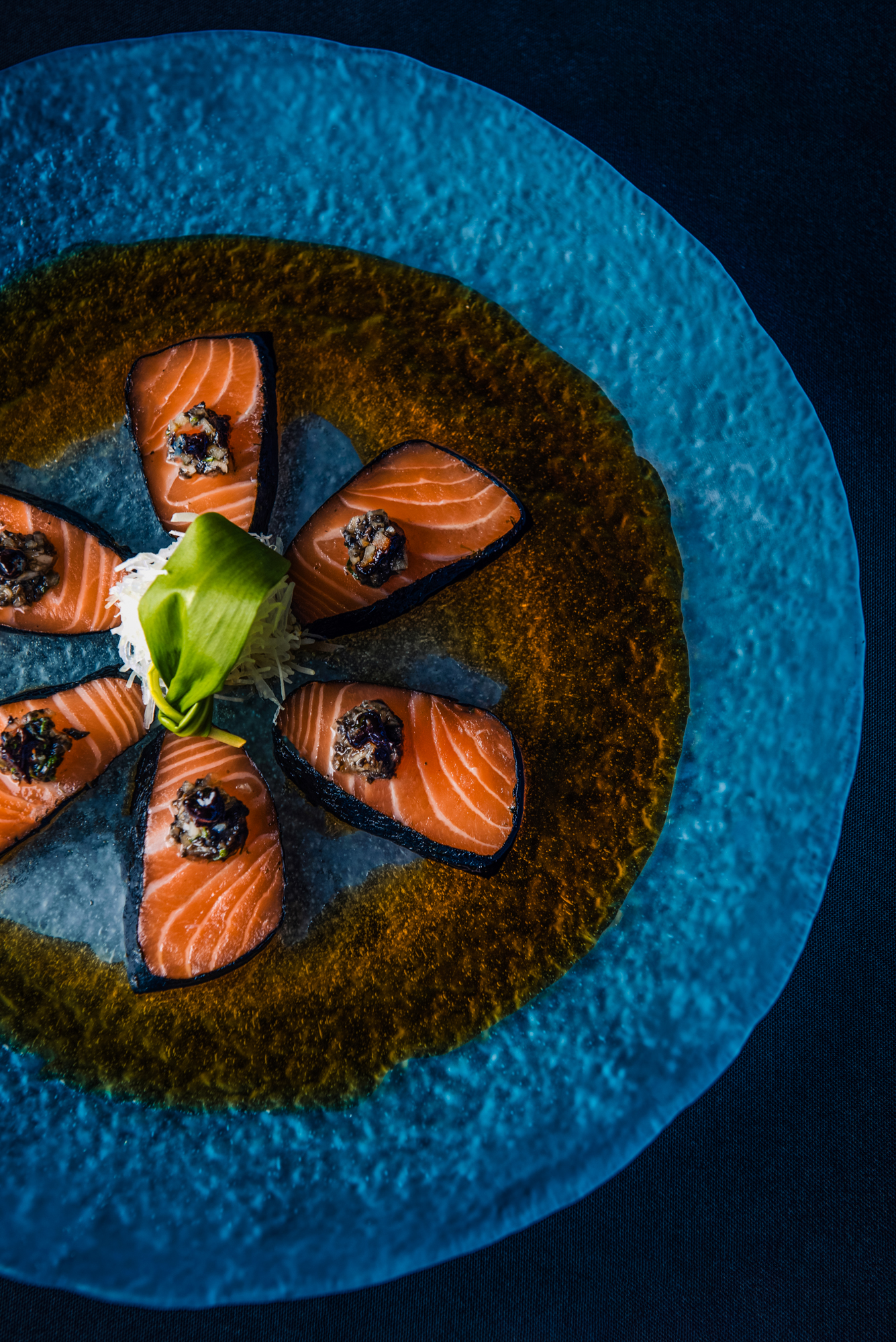 Close-up of salmon sashimi garnished with a savoury topping and green leaf centrepiece.