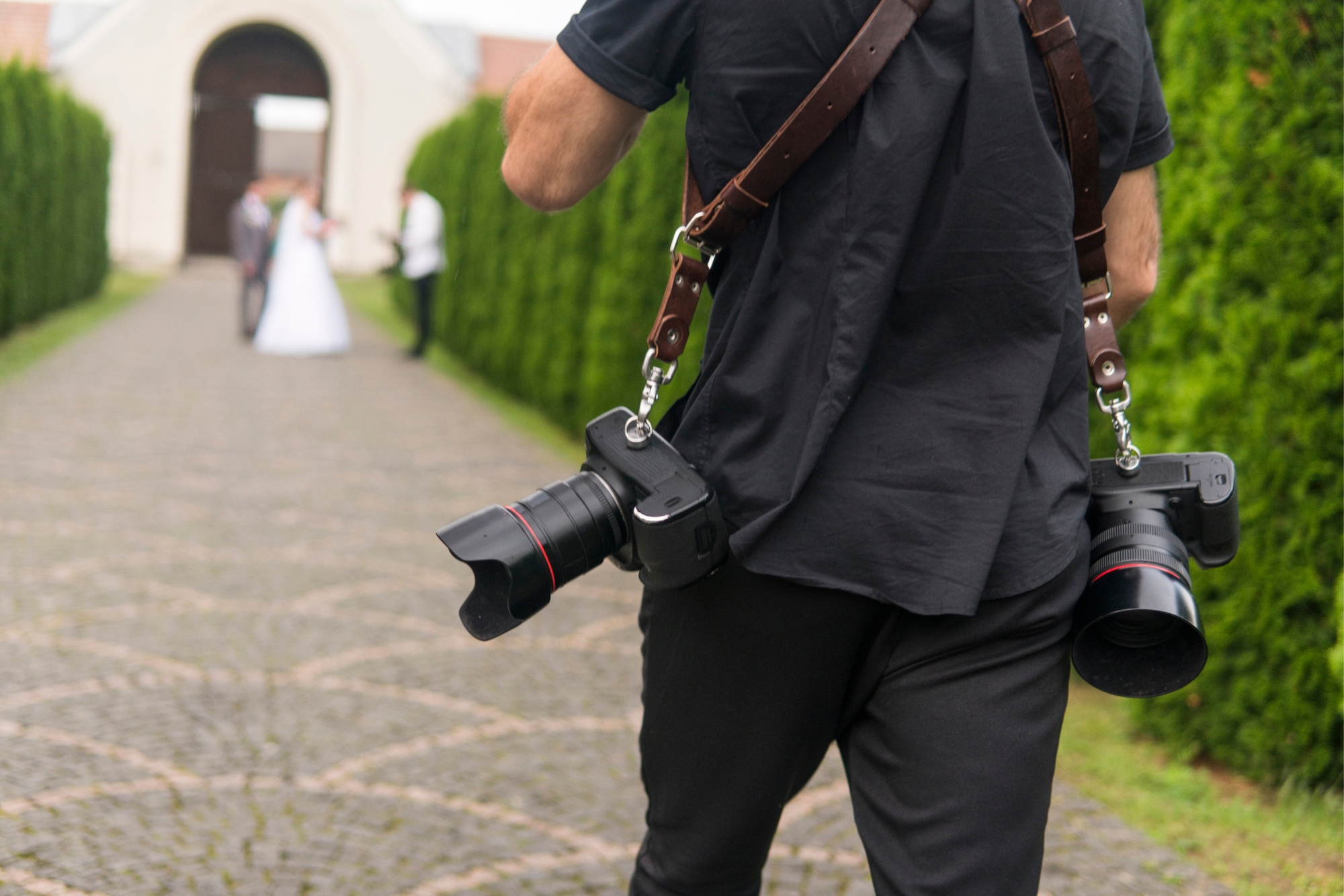 Wedding photographer carrying two cameras while walking towards a couple during an outdoor session.
