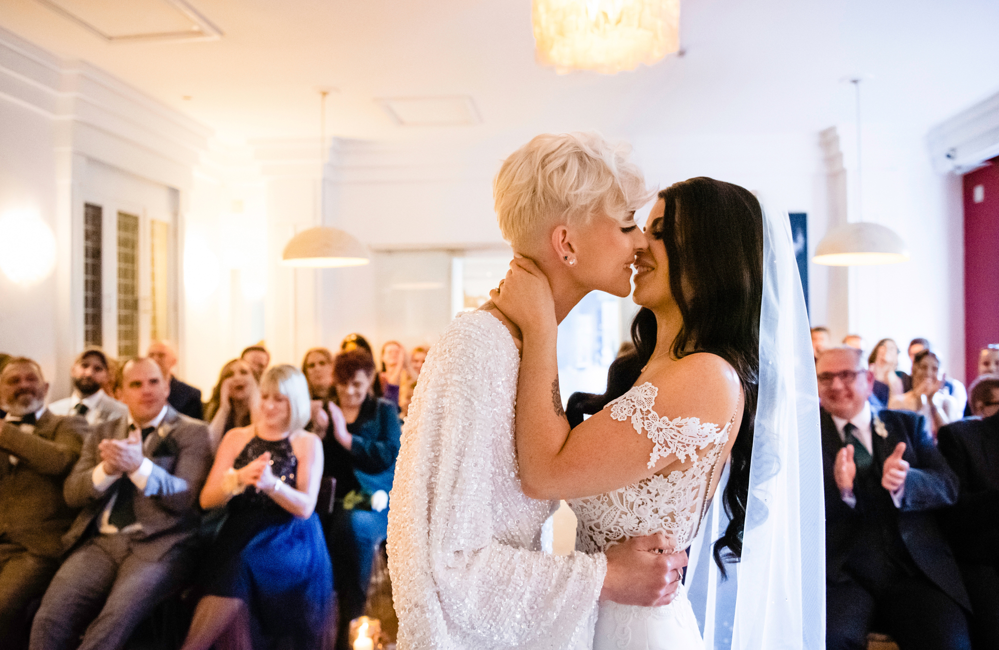 Brides sharing their first kiss at The Square Club Bristol while guests cheer and clap.