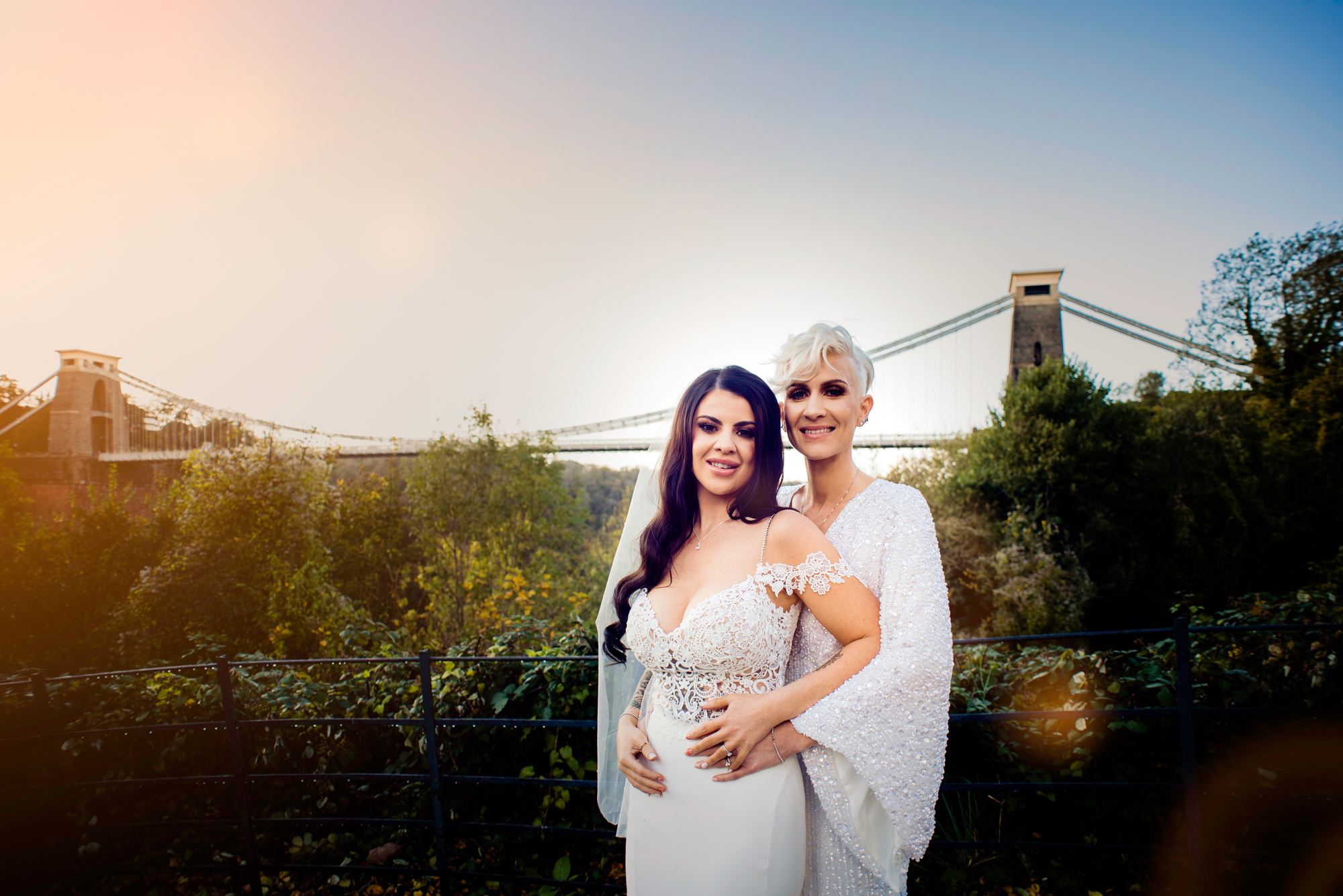 Brides embracing with the Clifton Suspension Bridge in the background, soft light over the gorge.