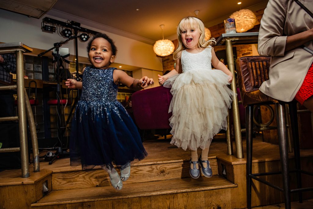 Two young flower girls in sparkly dresses jumping off a step and laughing during the reception.