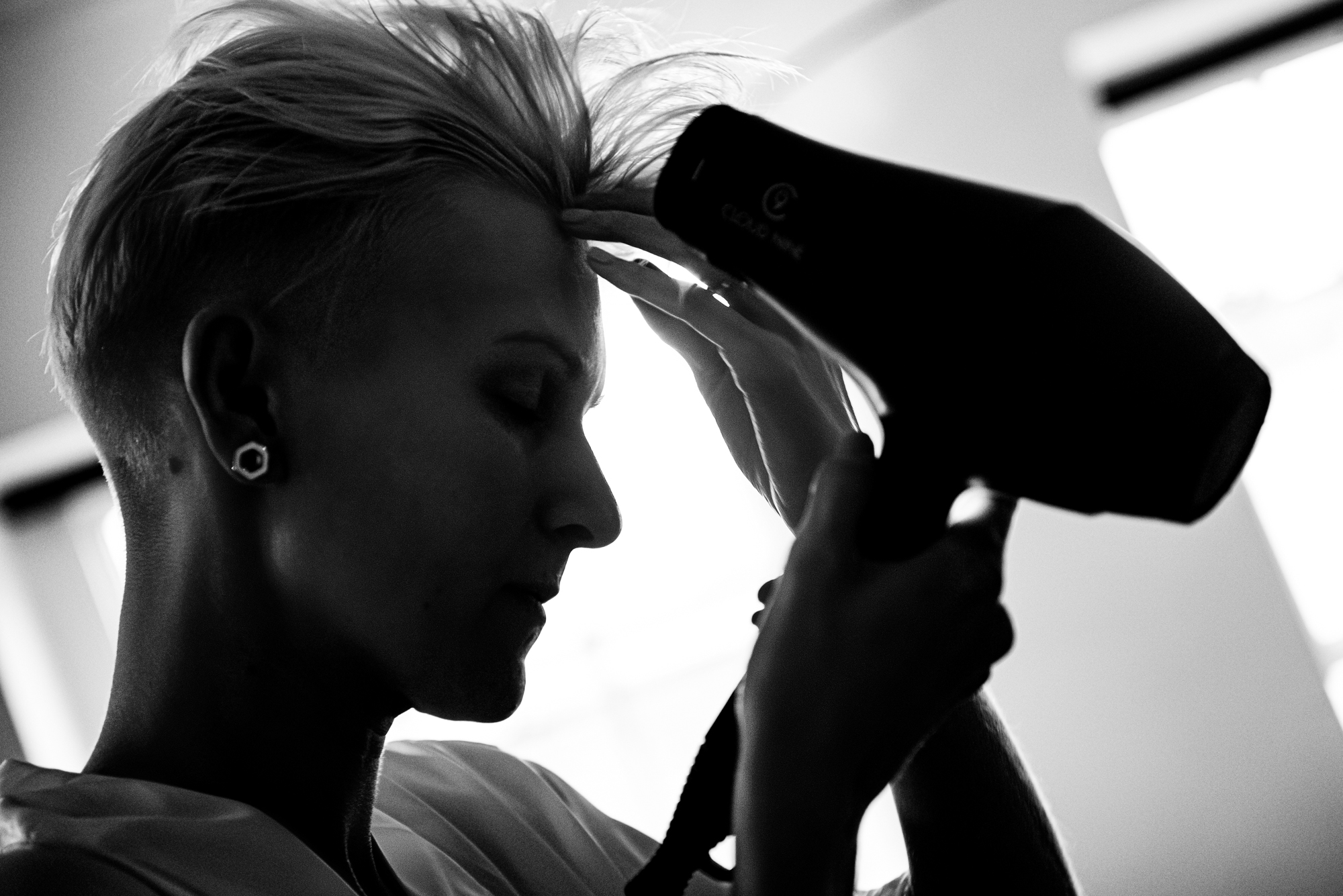 Bride blow-drying short blonde hair while getting ready in the morning light.