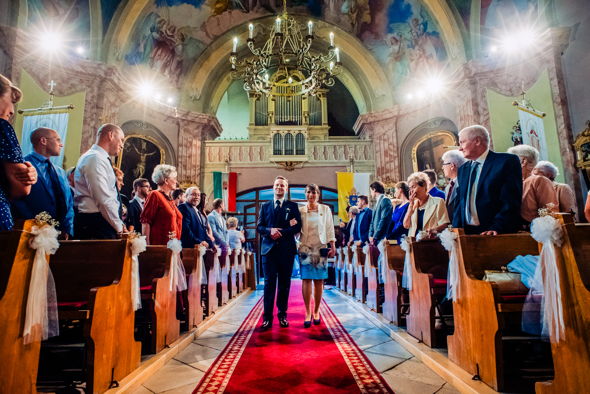 Groom walking down the church aisle with his mother during a traditional Hungarian wedding ceremony.