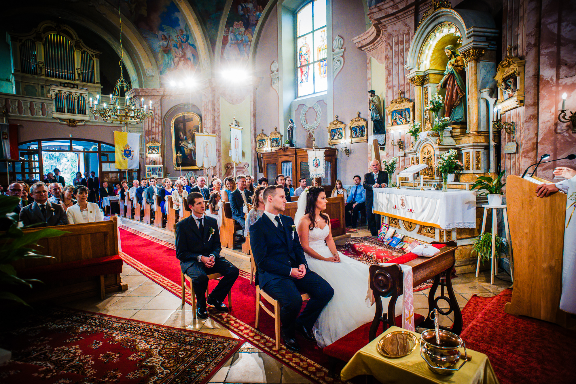 Wide view of a Catholic wedding ceremony inside an ornate church in Hungary with seated guests.