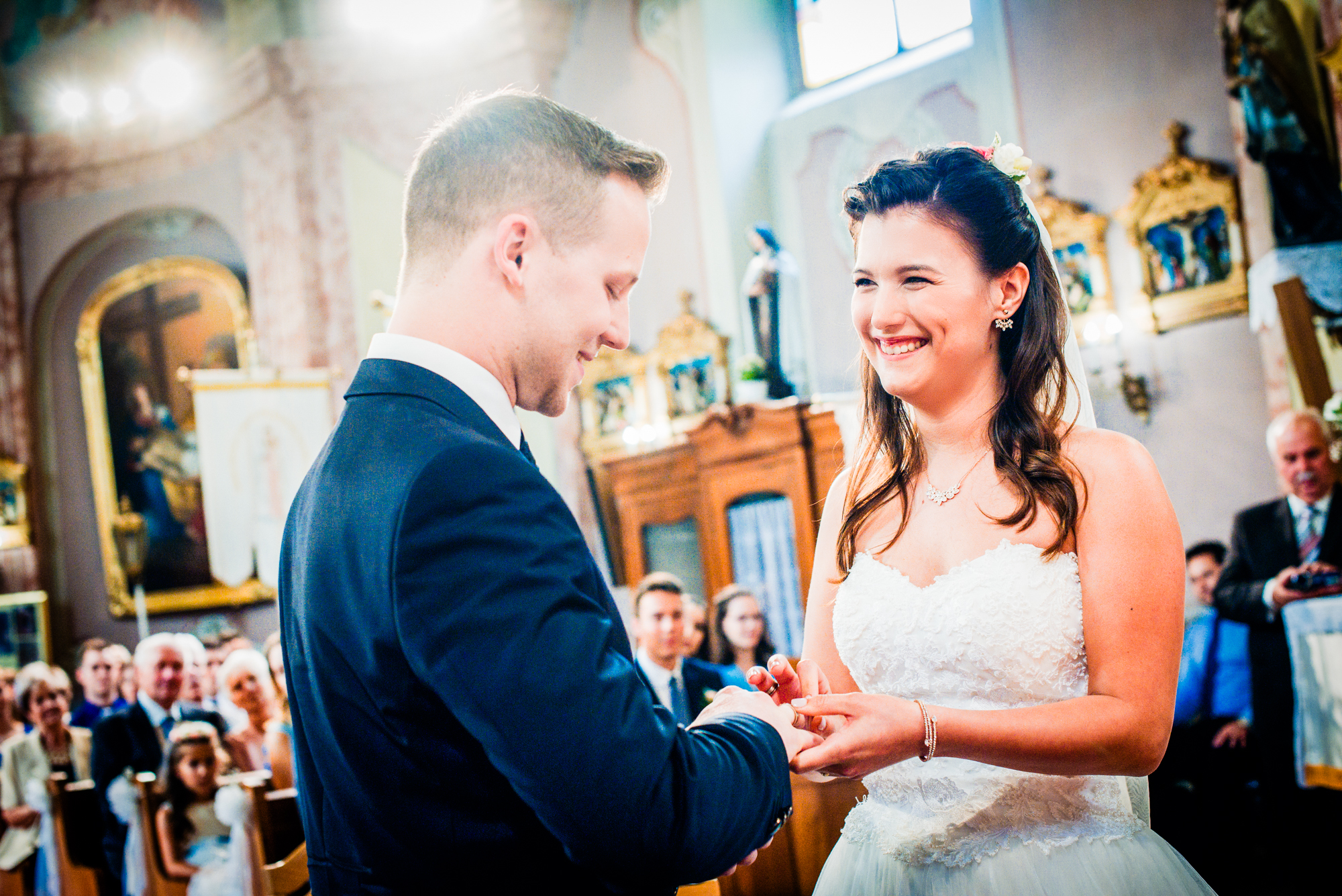 Bride smiling while placing a wedding ring on the groom’s finger during church ceremony.