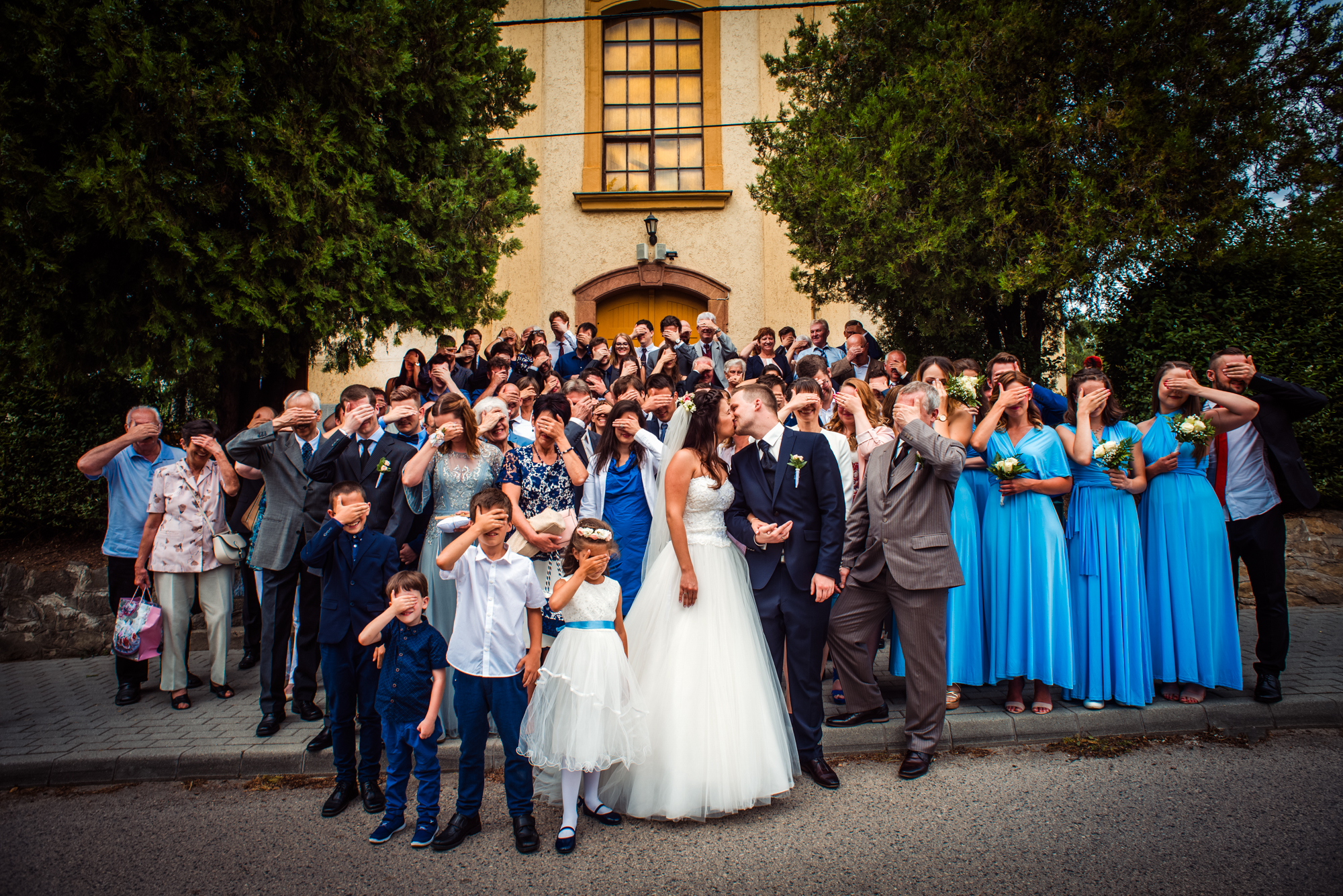 Bride and groom kissing during a playful wedding group photo outside the church.
