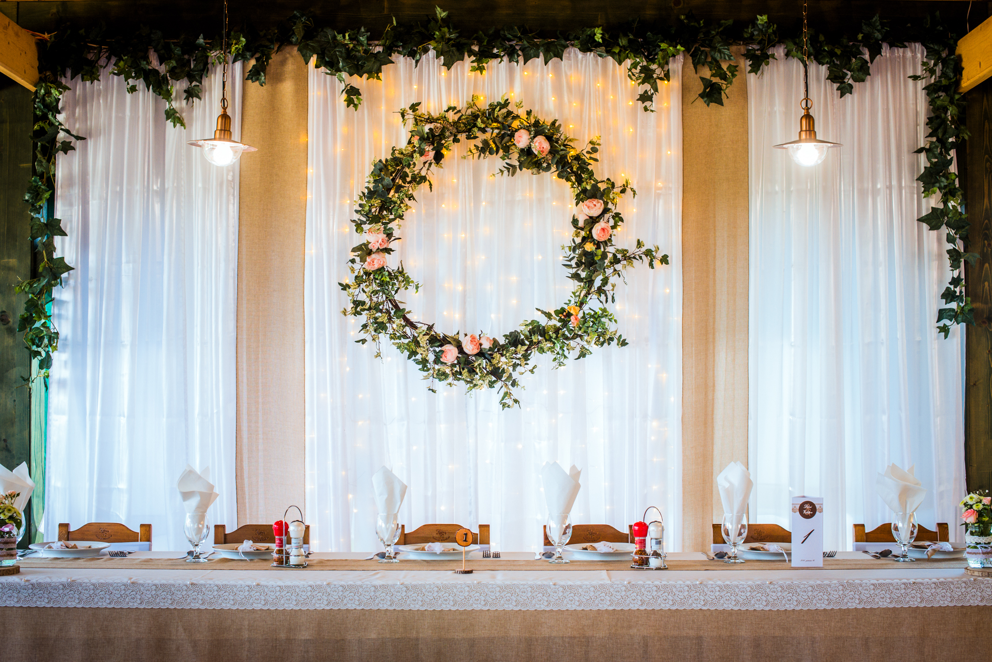 Rustic wedding head table with floral wreath backdrop and warm fairy lights at vineyard reception.