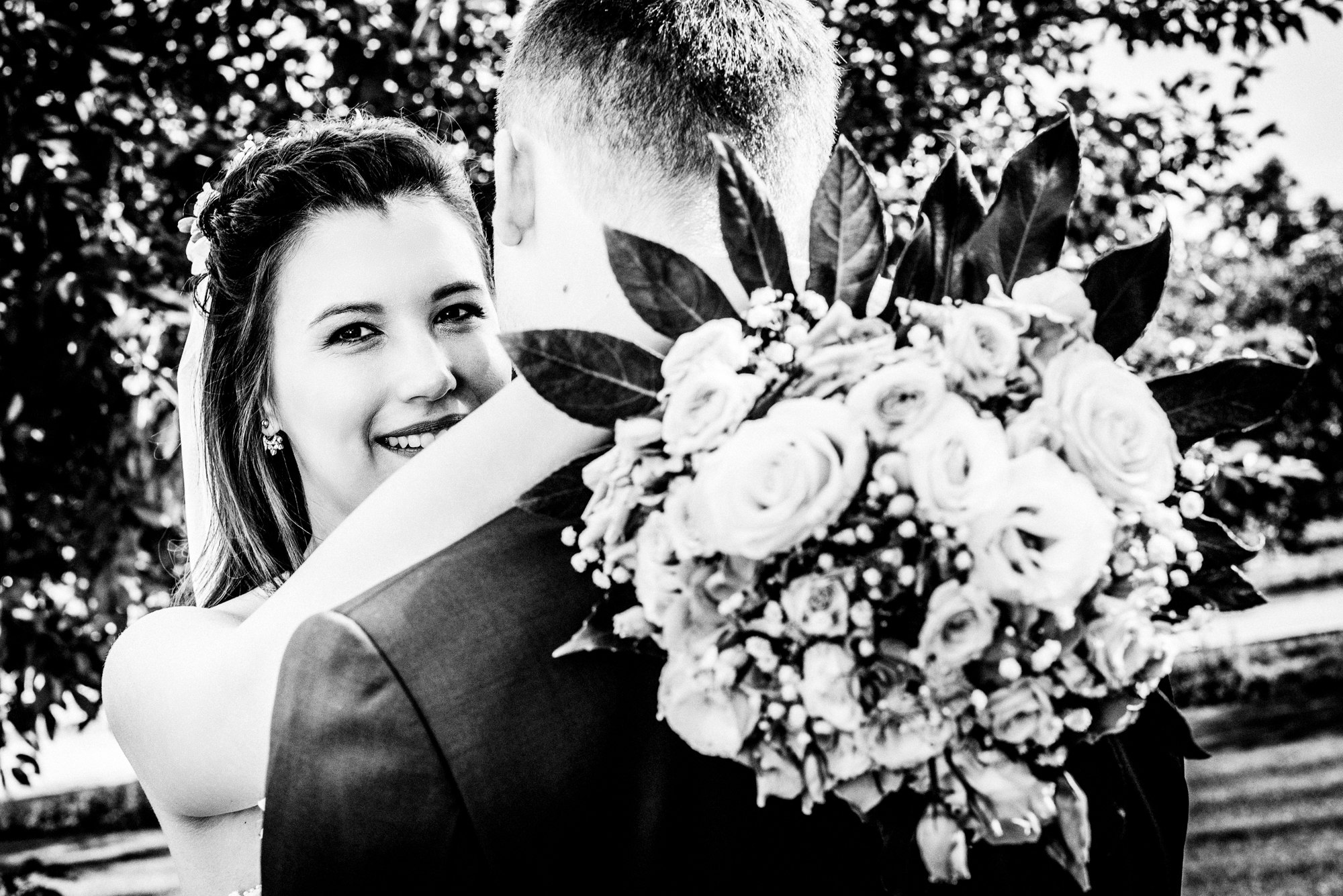 Bride smiling over groom’s shoulder while holding bouquet during outdoor Hungary vineyard wedding.