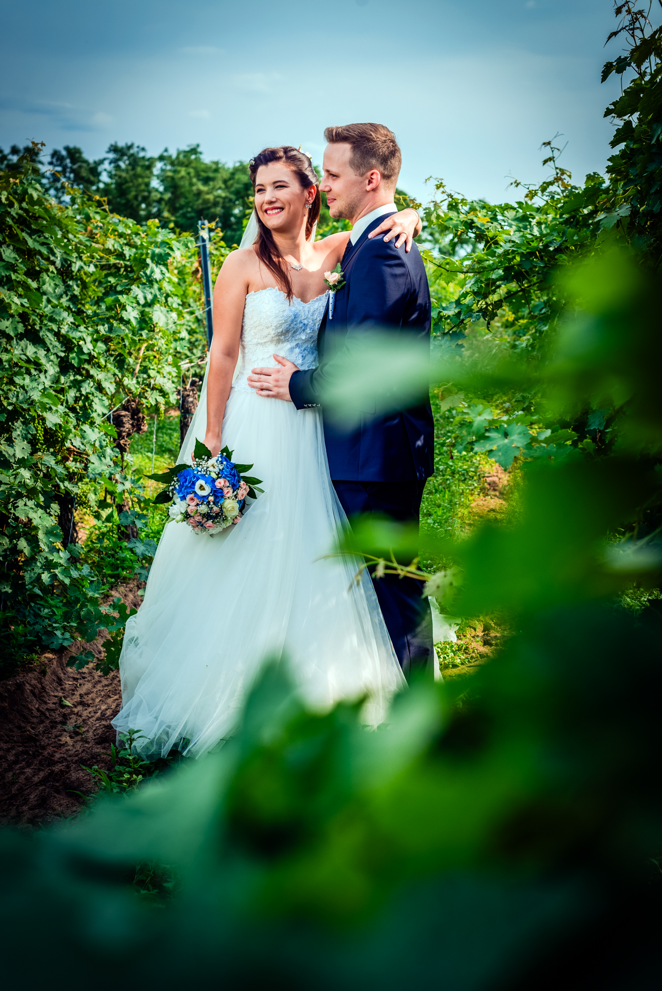 Bride and groom standing together in vineyard with blue and pink wedding bouquet in Hungary.