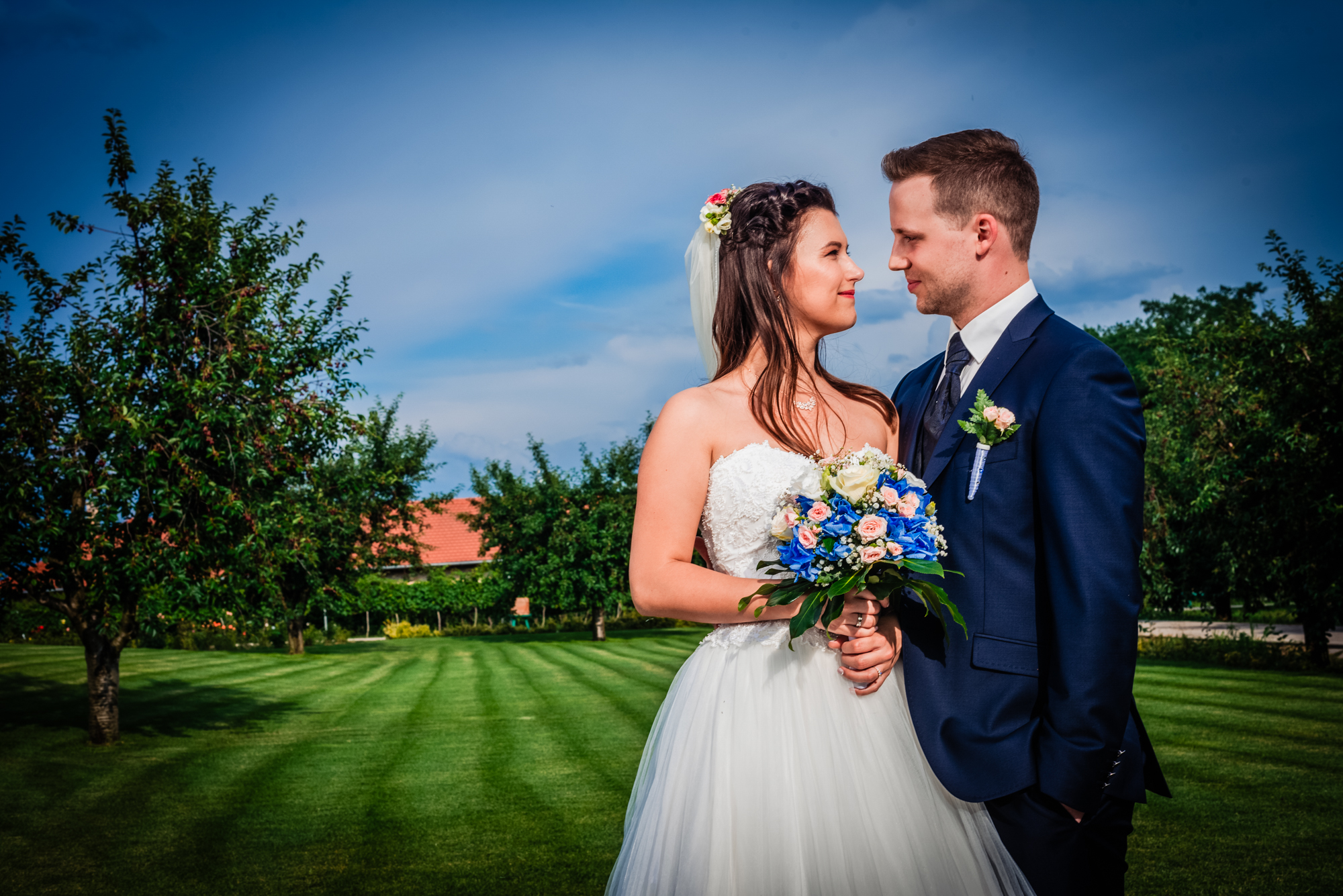 Bride smiling while groom embraces her through sunlit vineyard leaves at outdoor Hungary wedding.