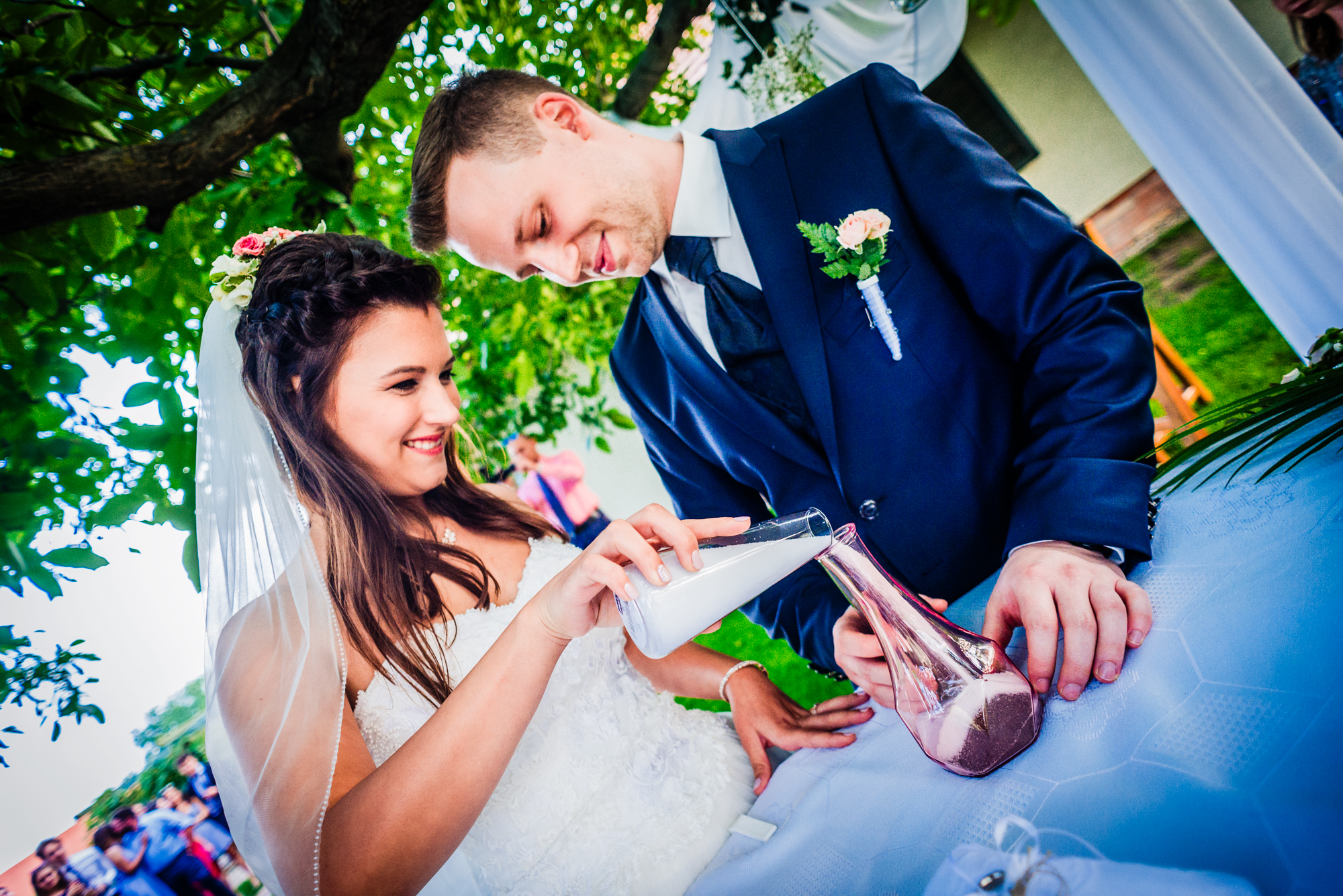 Bride pouring ceremonial sand with groom during outdoor wedding ritual beneath trees in Hungary.