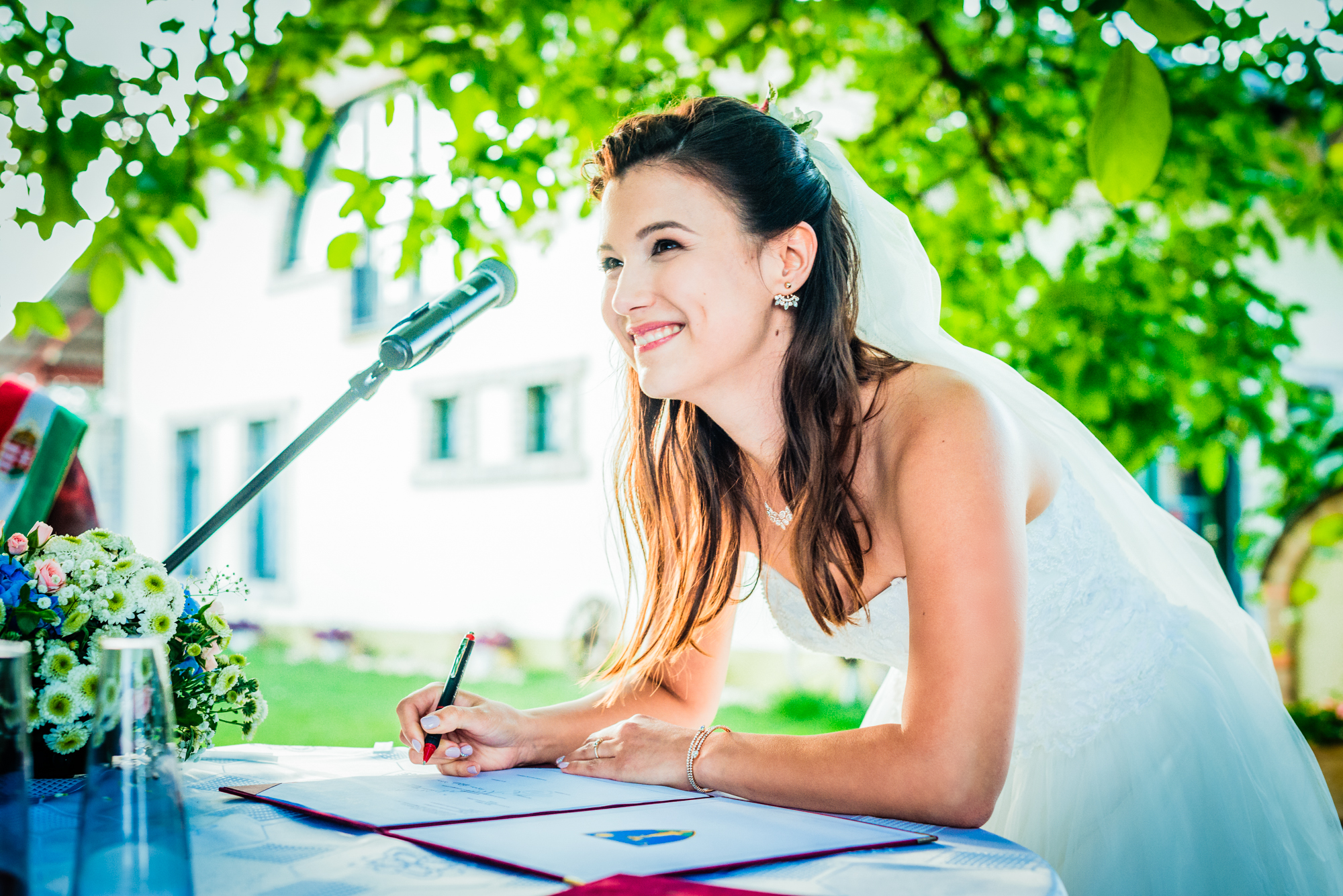 Bride signing wedding documents outdoors beneath trees during summer ceremony in Hungary.