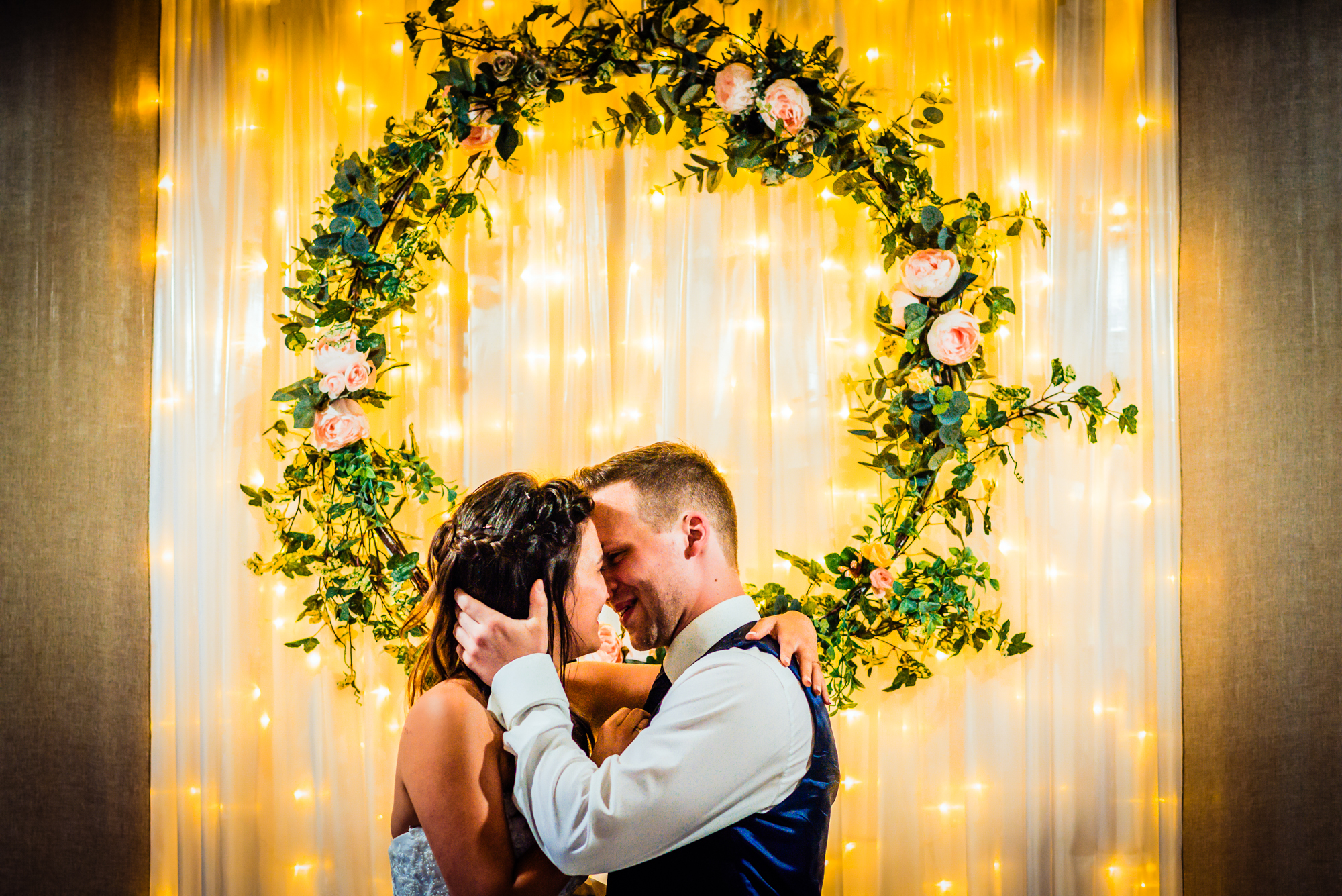 Bride and groom embracing beneath illuminated floral wreath during evening wedding reception dance.