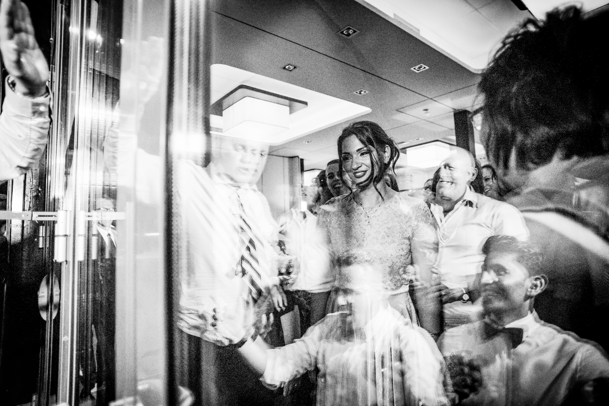 Moment of the bride with reflections of the groom in the window and guests.