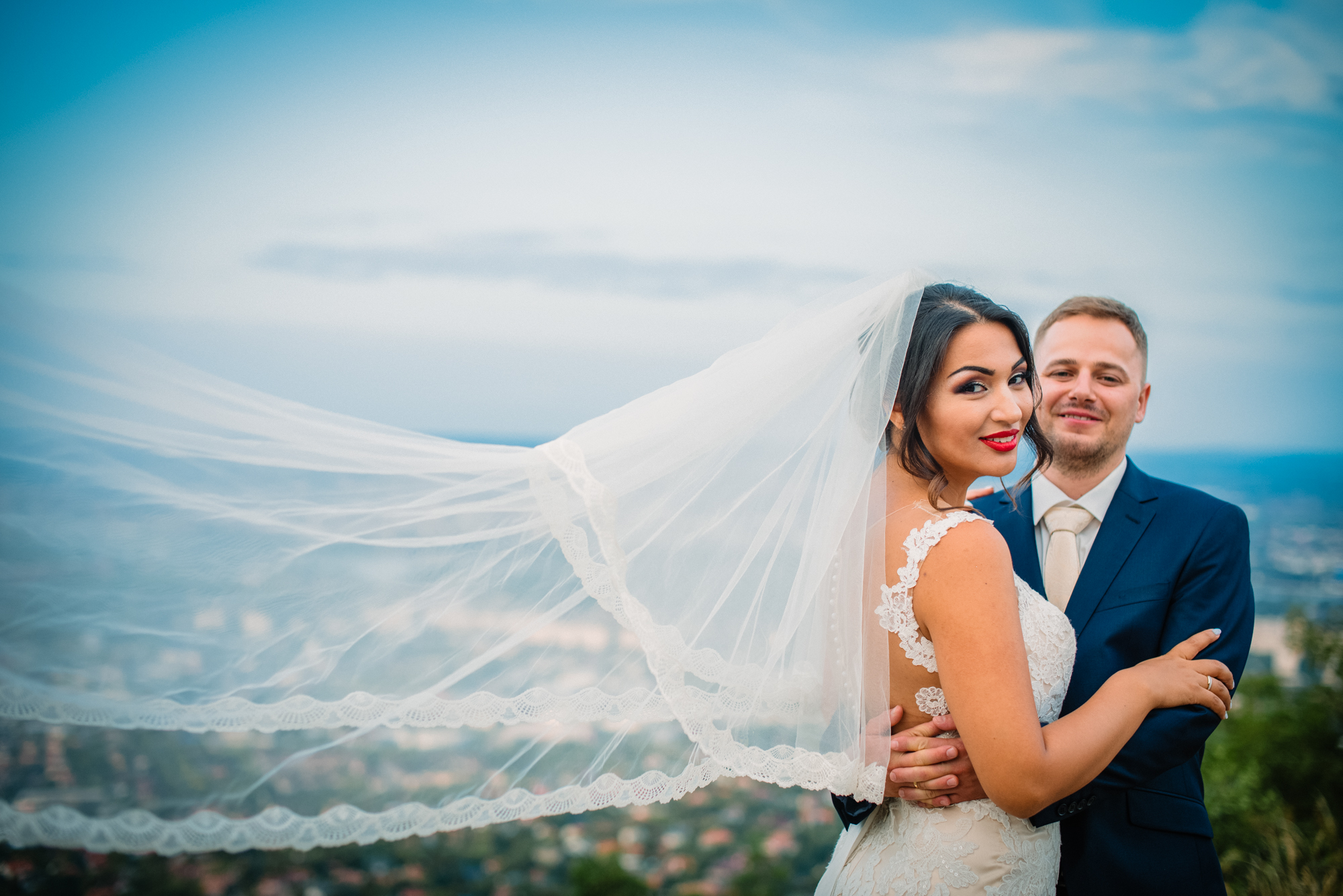 Bride smiling at camera with veil blowing in wind while groom holds her during sunset portrait.