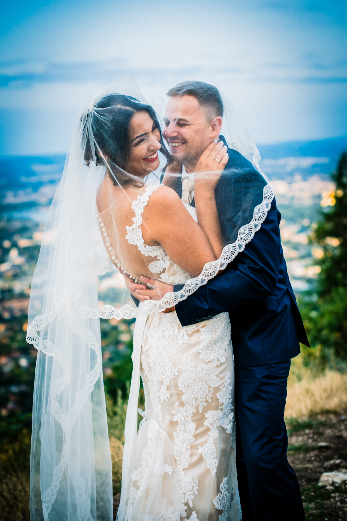 Bride and groom laughing closely together wrapped in veil with blurred Budapest skyline behind them.