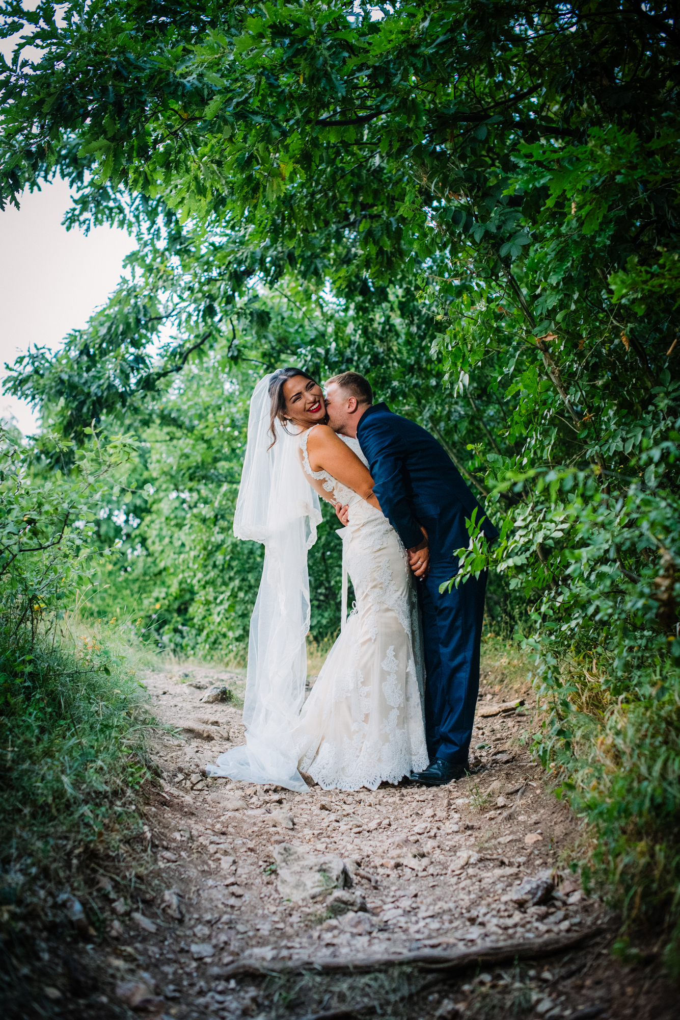 Groom kissing bride’s cheek on forest path during relaxed and playful wedding portrait session.