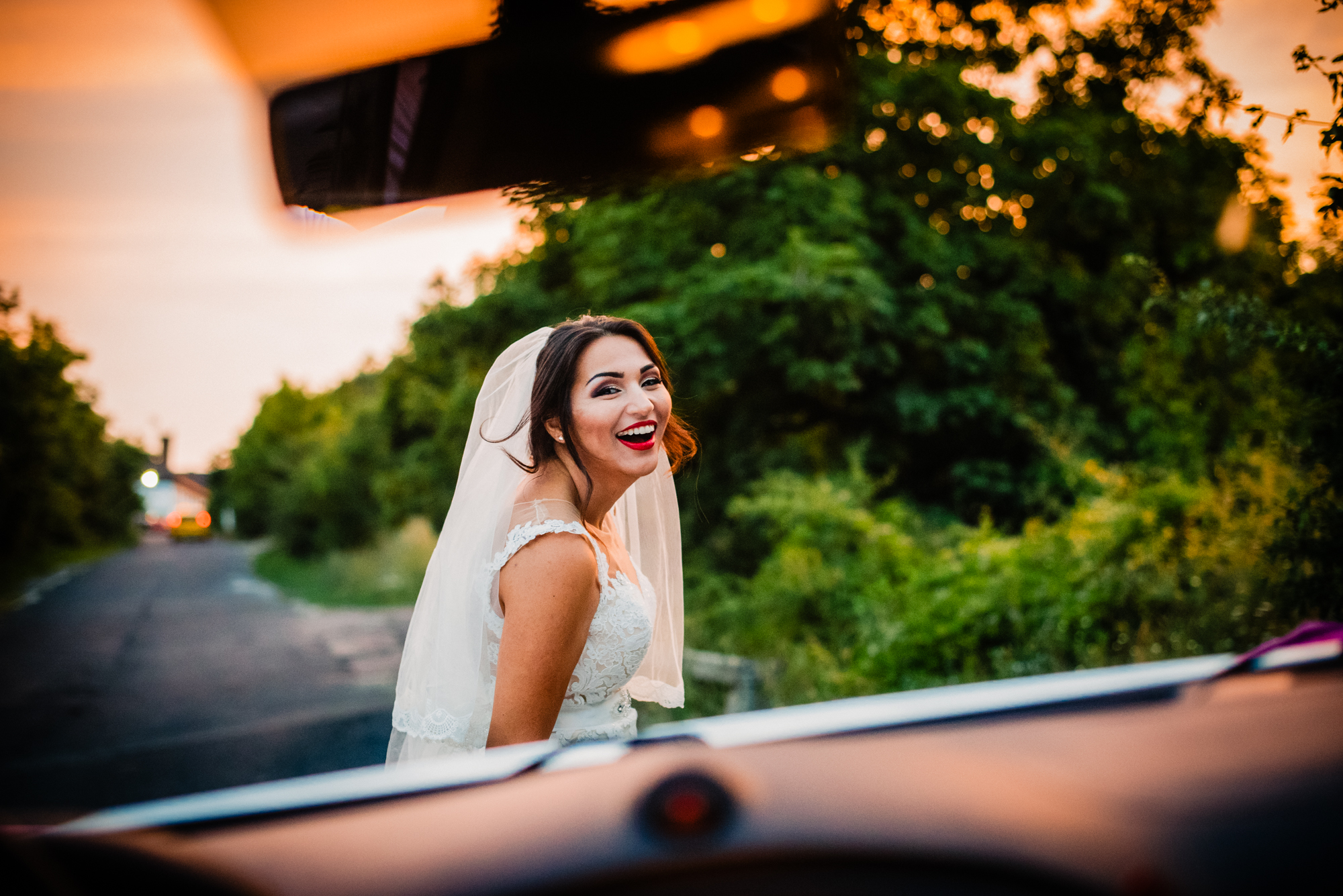 Bride smiling brightly captured through car window reflection during candid wedding moment.
