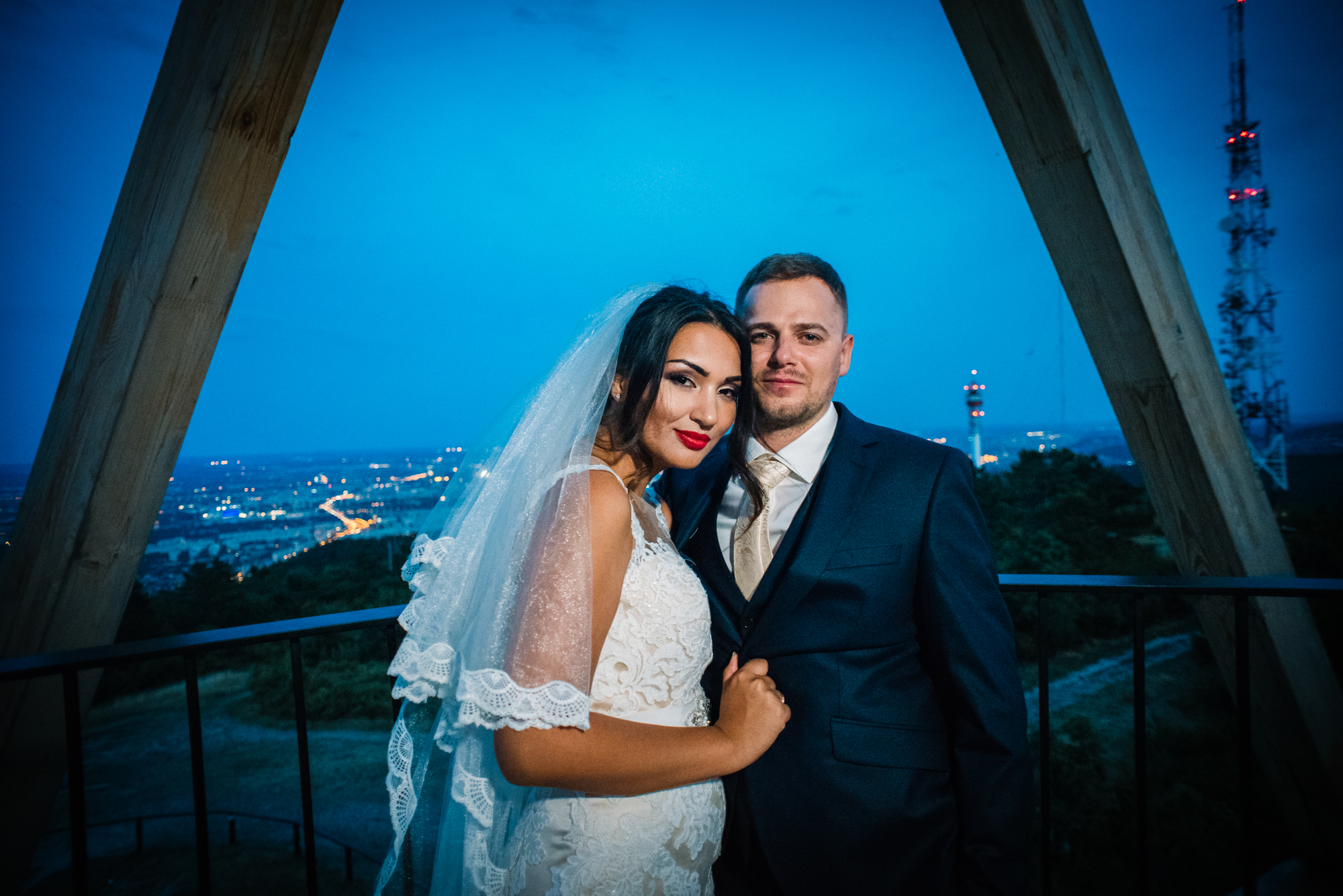 Bride and groom standing under wooden structure during blue hour with Budapest city lights behind them.