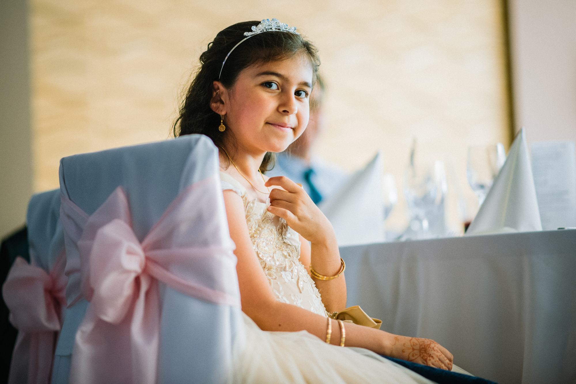 Young girl in white dress with tiara sitting at reception table, soft candid portrait.